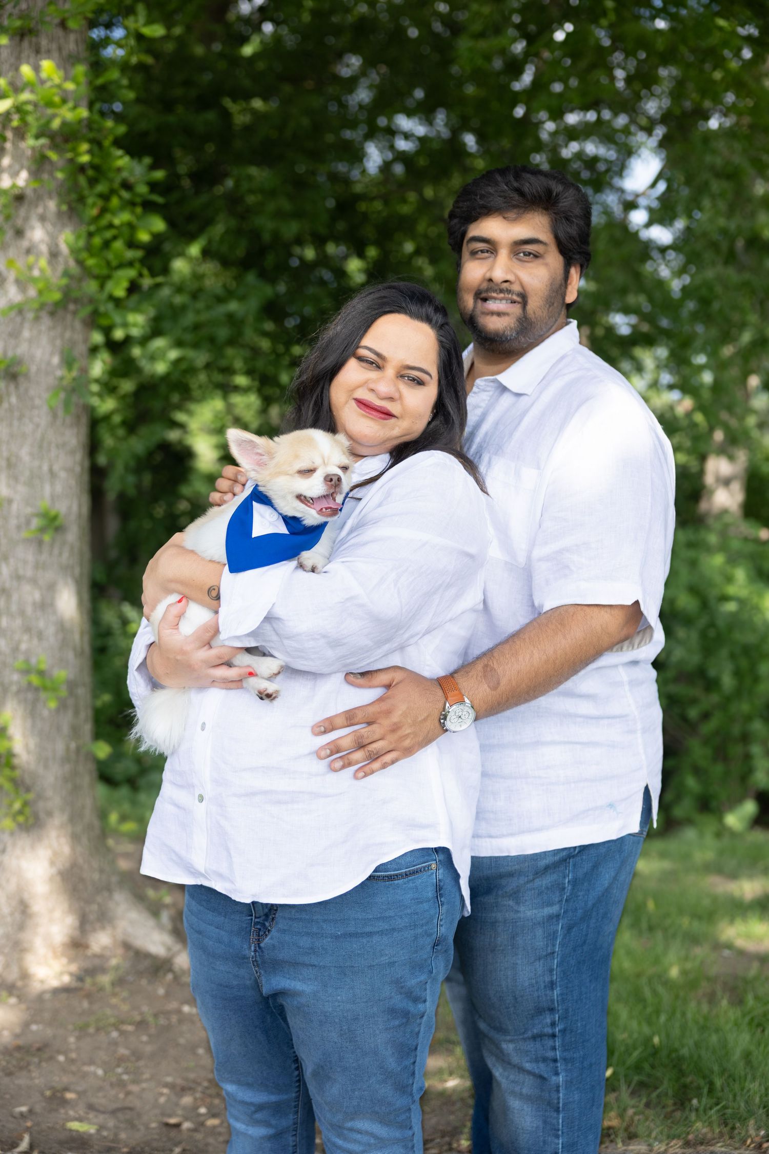Smiling couple in white shirts holding a small Chihuahua dog dressed in blue outdoors.