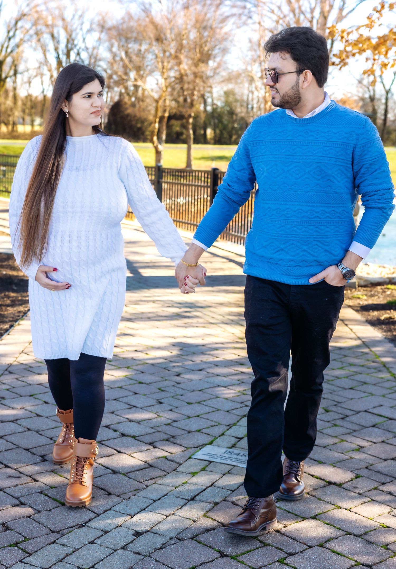 Expecting couple walking hand in hand on brick path in sunny autumn park.