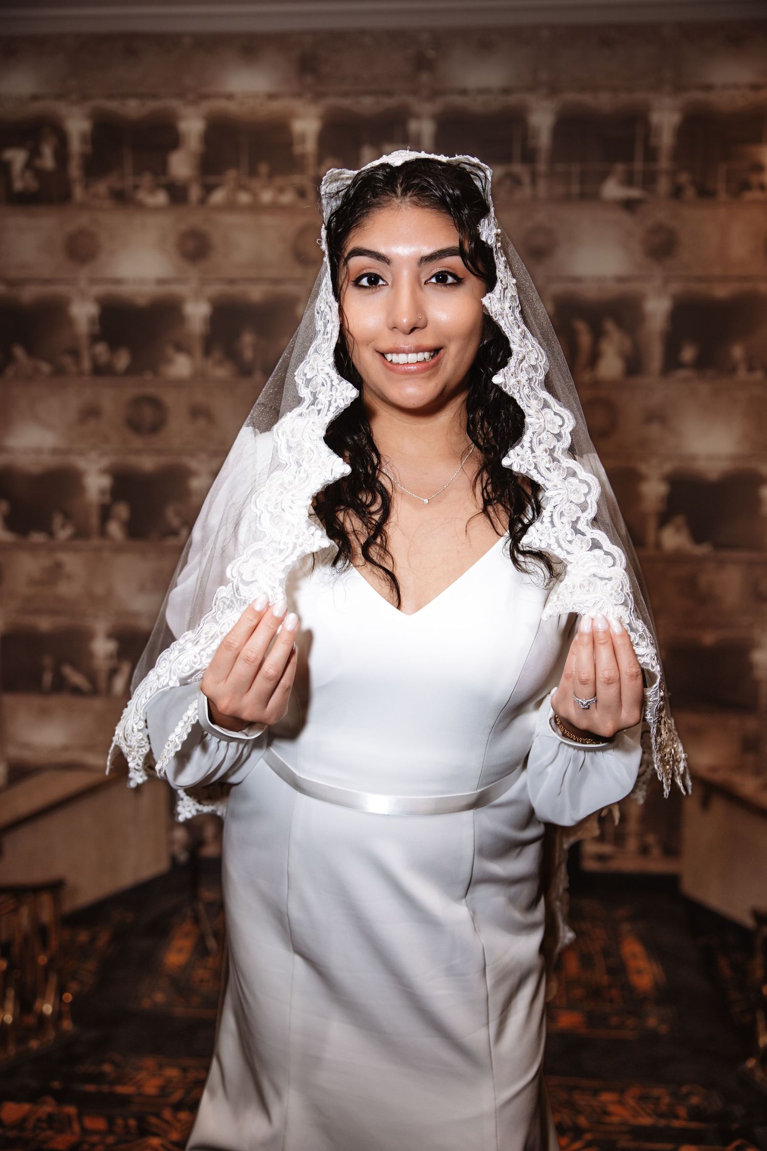 Smiling bride in white dress and lace mantilla veil gestures with hands in decorated wedding venue.