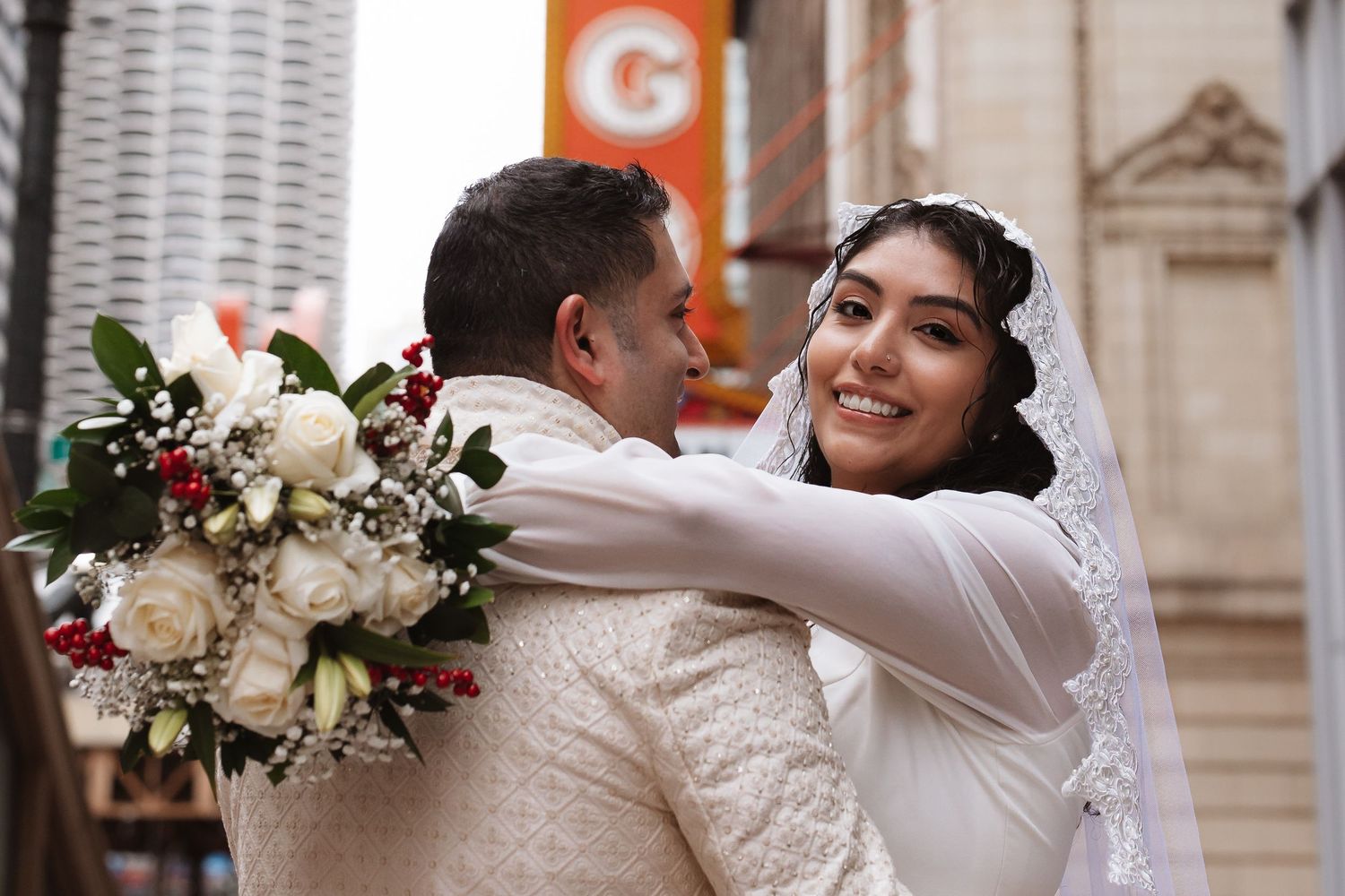 Close-up of bride smiling while hugging groom holding white and red wedding bouquet.
