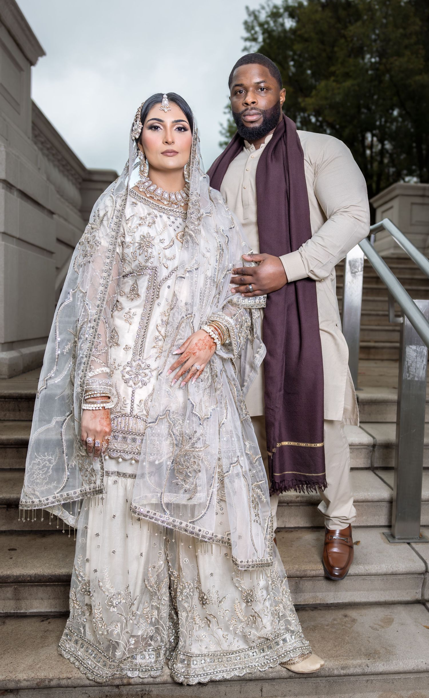 Bride in ornate white gharara with gold embroidery and groom in beige kurta on outdoor stairs.