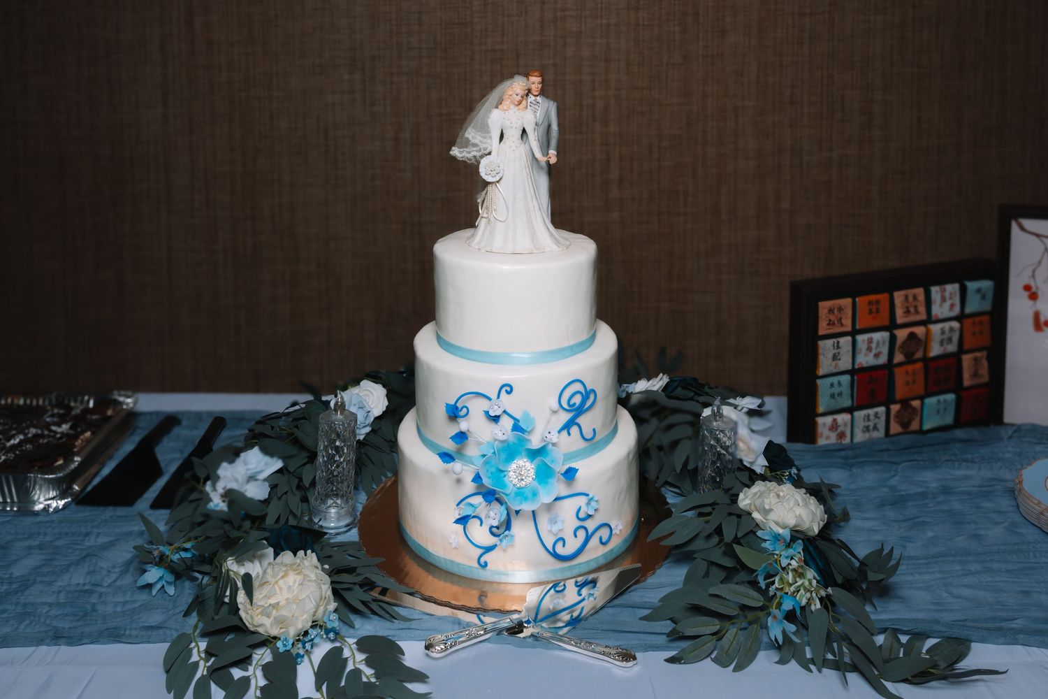 Three-tier white wedding cake with blue floral details and bride and groom topper on decorated table.