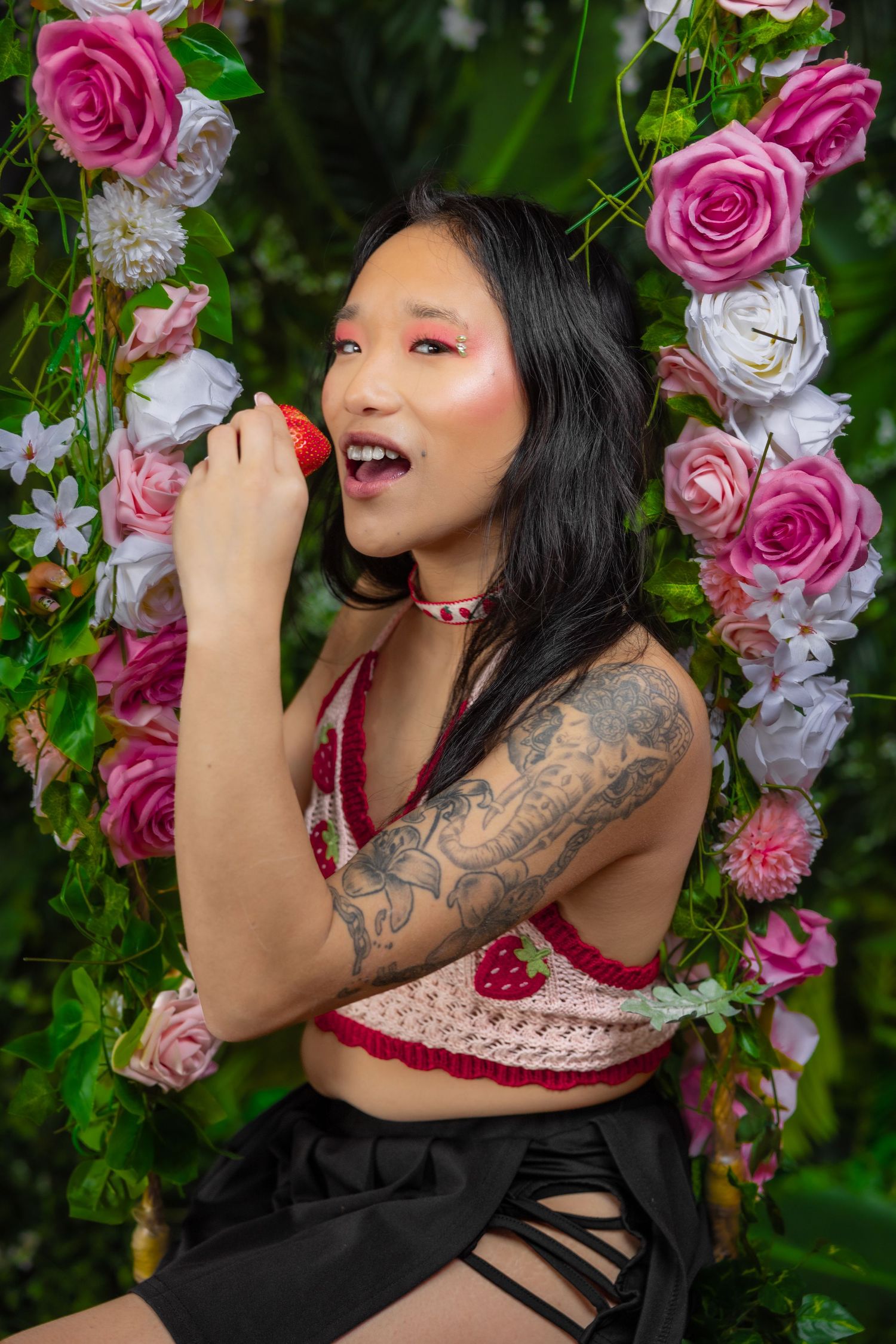 Tattooed Asian woman in crochet crop top eats strawberry on floral rose swing.