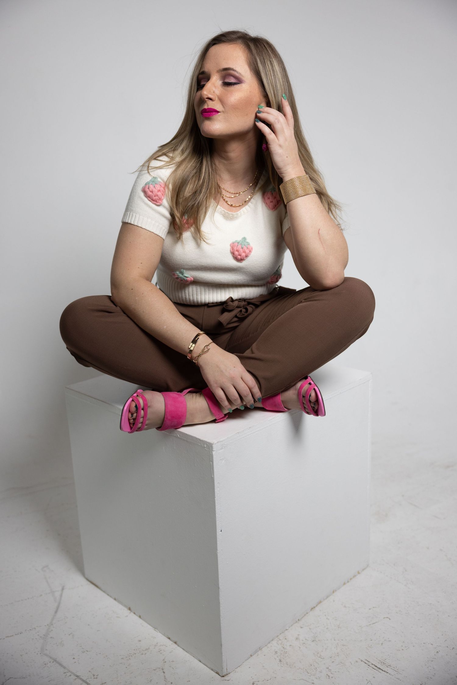 Blonde woman in strawberry print top and brown pants sitting cross-legged on white cube in studio.