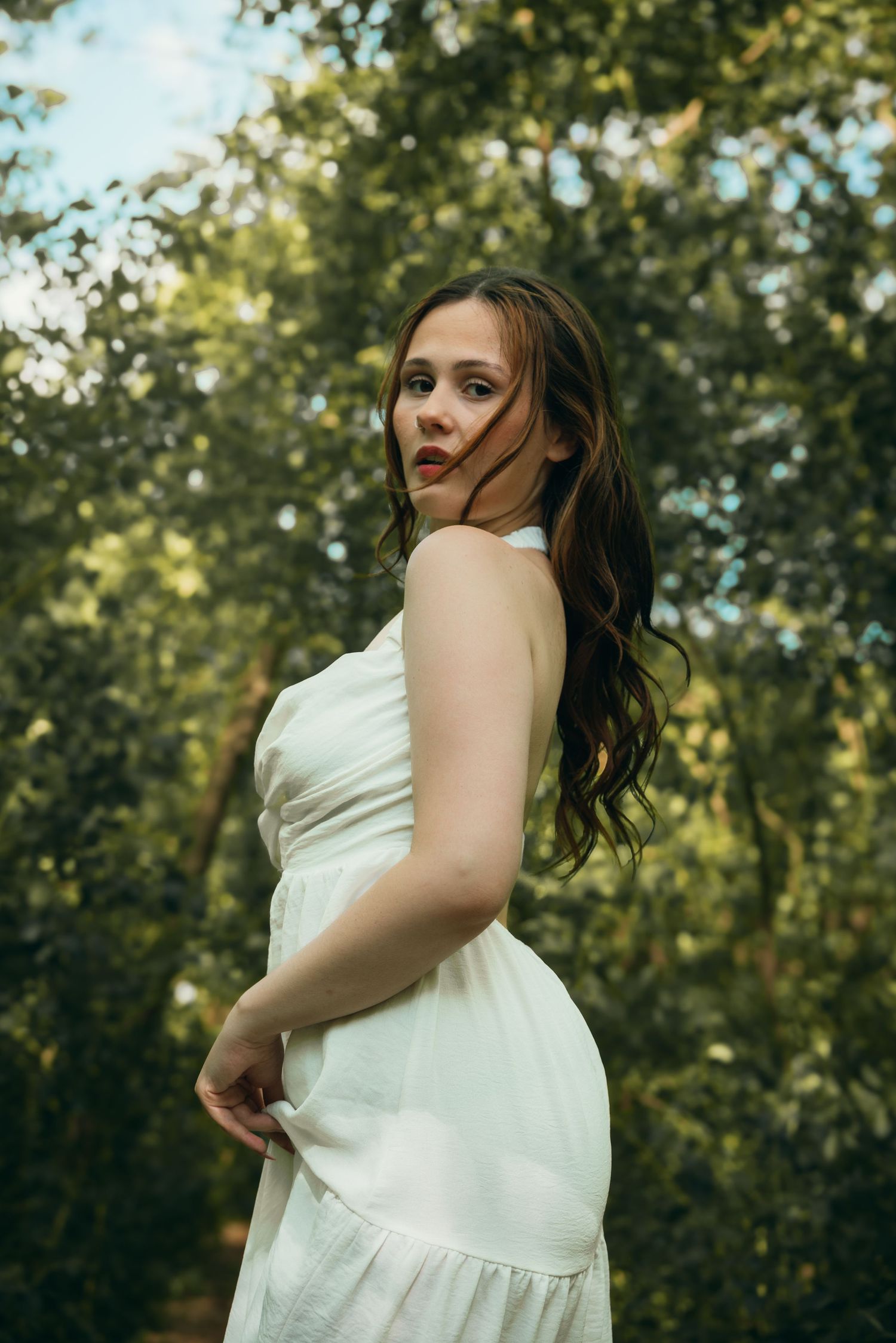 A brunette woman in a white sleeveless dress poses gracefully among vibrant green trees in a forest.