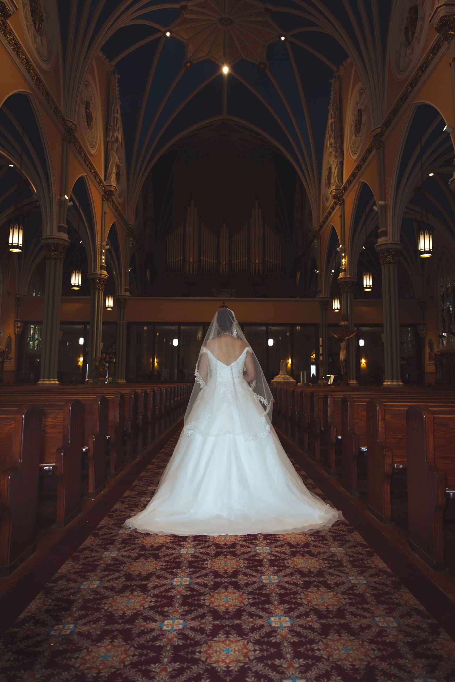 Bride in white ball gown and veil standing alone in ornate Gothic cathedral aisle from behind.