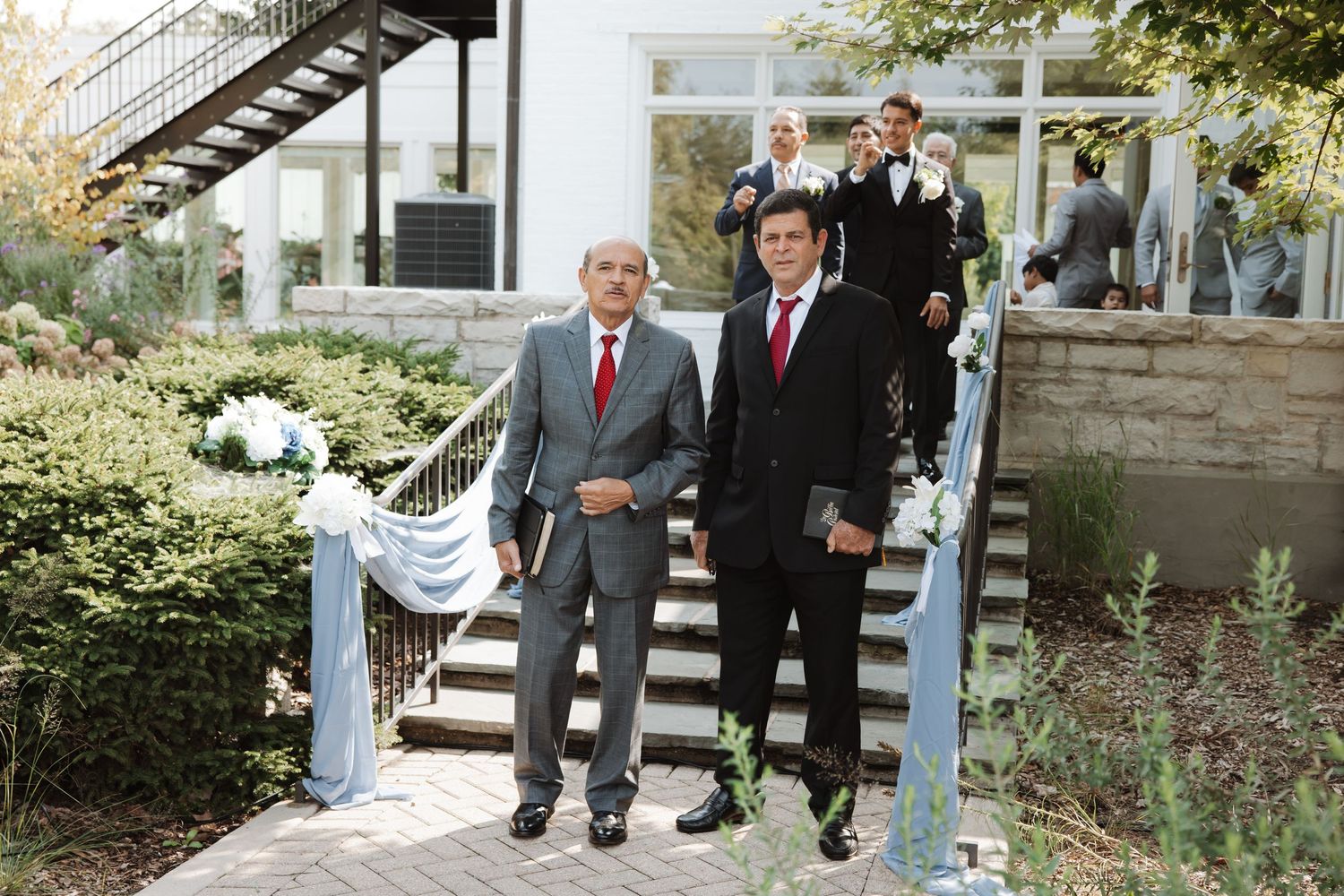 Two men in dark suits with red ties pose on outdoor steps at a wedding venue.