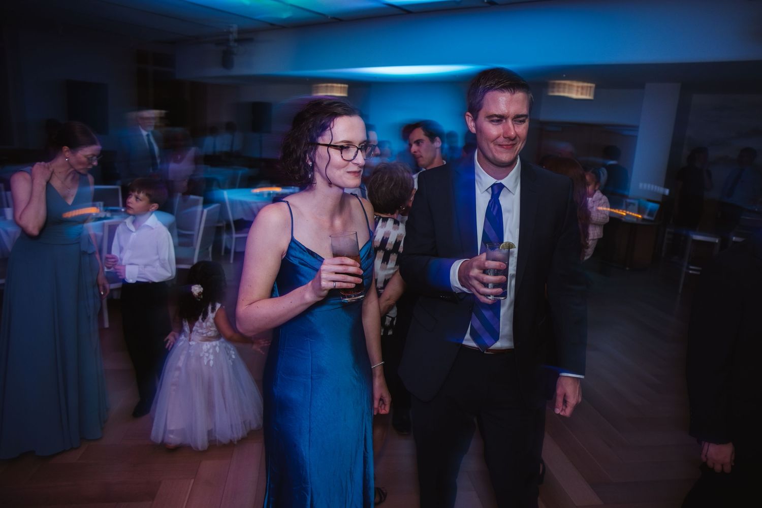 A smiling couple in formal attire holds drinks while enjoying a wedding reception dance floor with blue lighting.