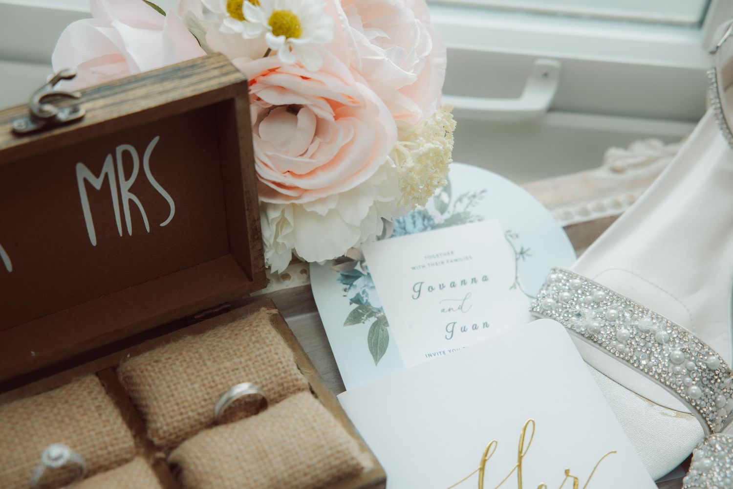 Wedding flat lay with blush roses, rings, MRS box, and floral invitation.
