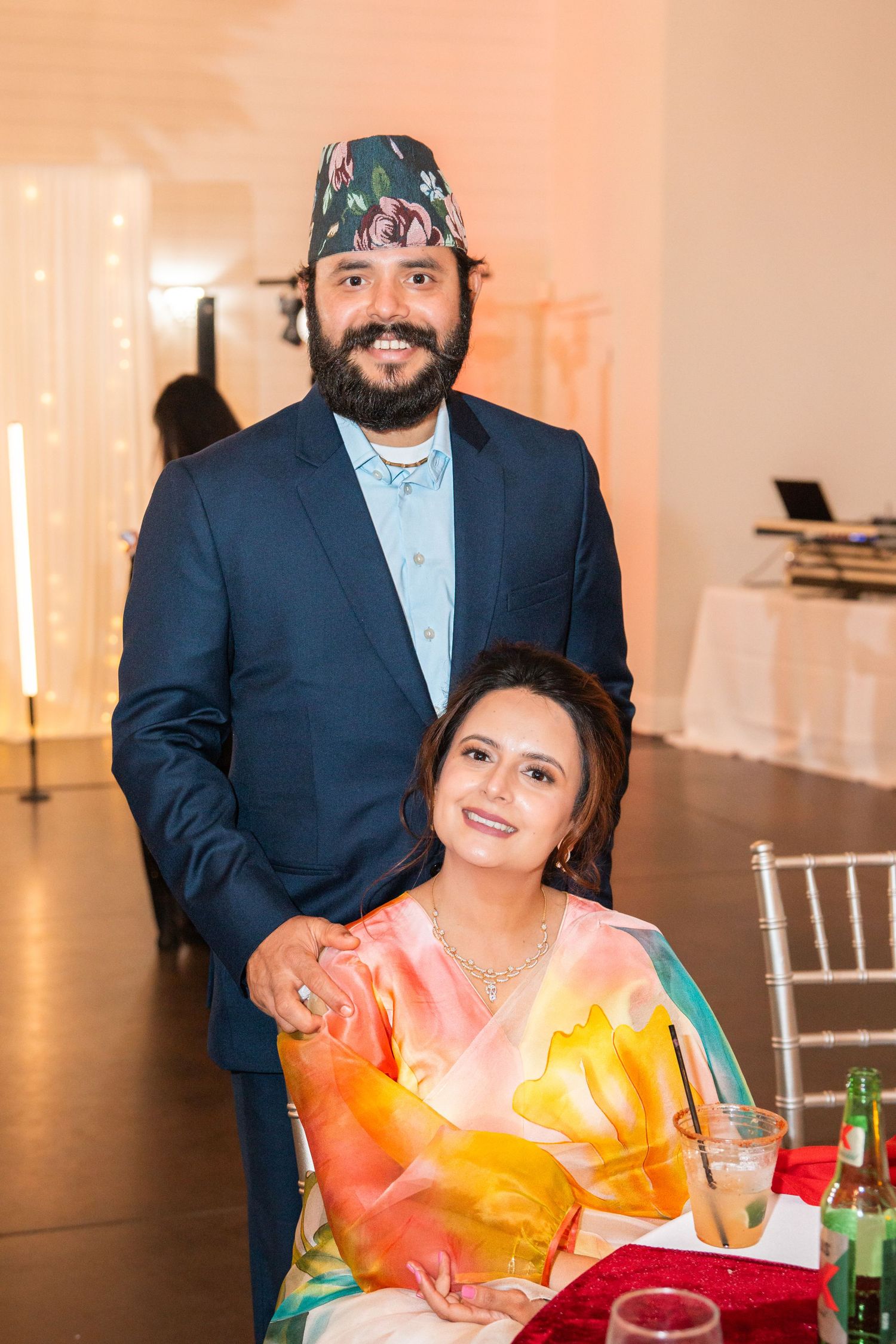 Couple posing at a South Asian wedding reception, man wearing traditional Nepali topi hat and blue suit.