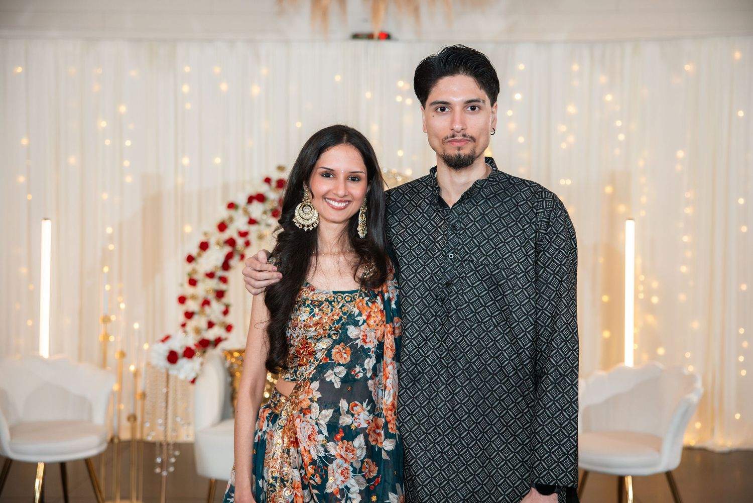 Smiling couple dressed in colorful Indian outfits standing before a floral and fairy light backdrop.
