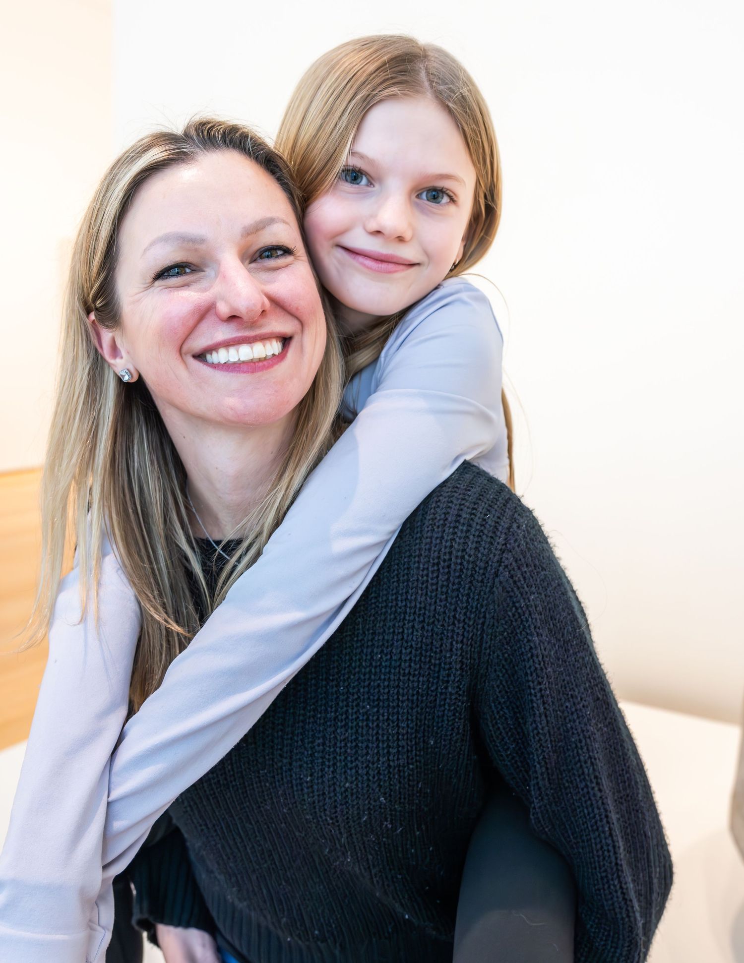 A woman smiles warmly as a young girl hugs her from behind in a bright indoor setting.
