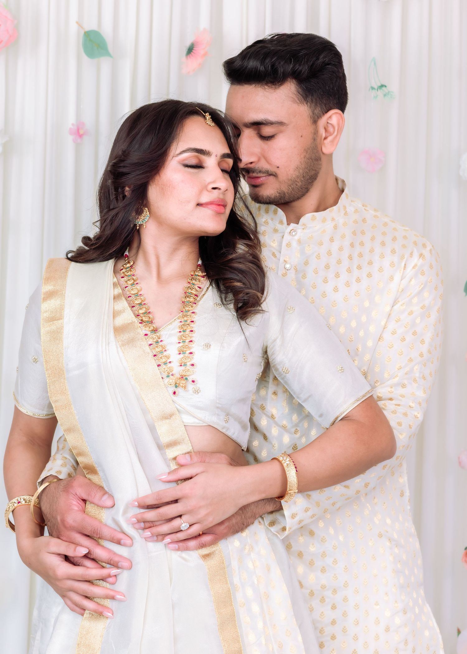 South Asian groom in white kurta embracing bride in white lehenga during romantic wedding photoshoot.