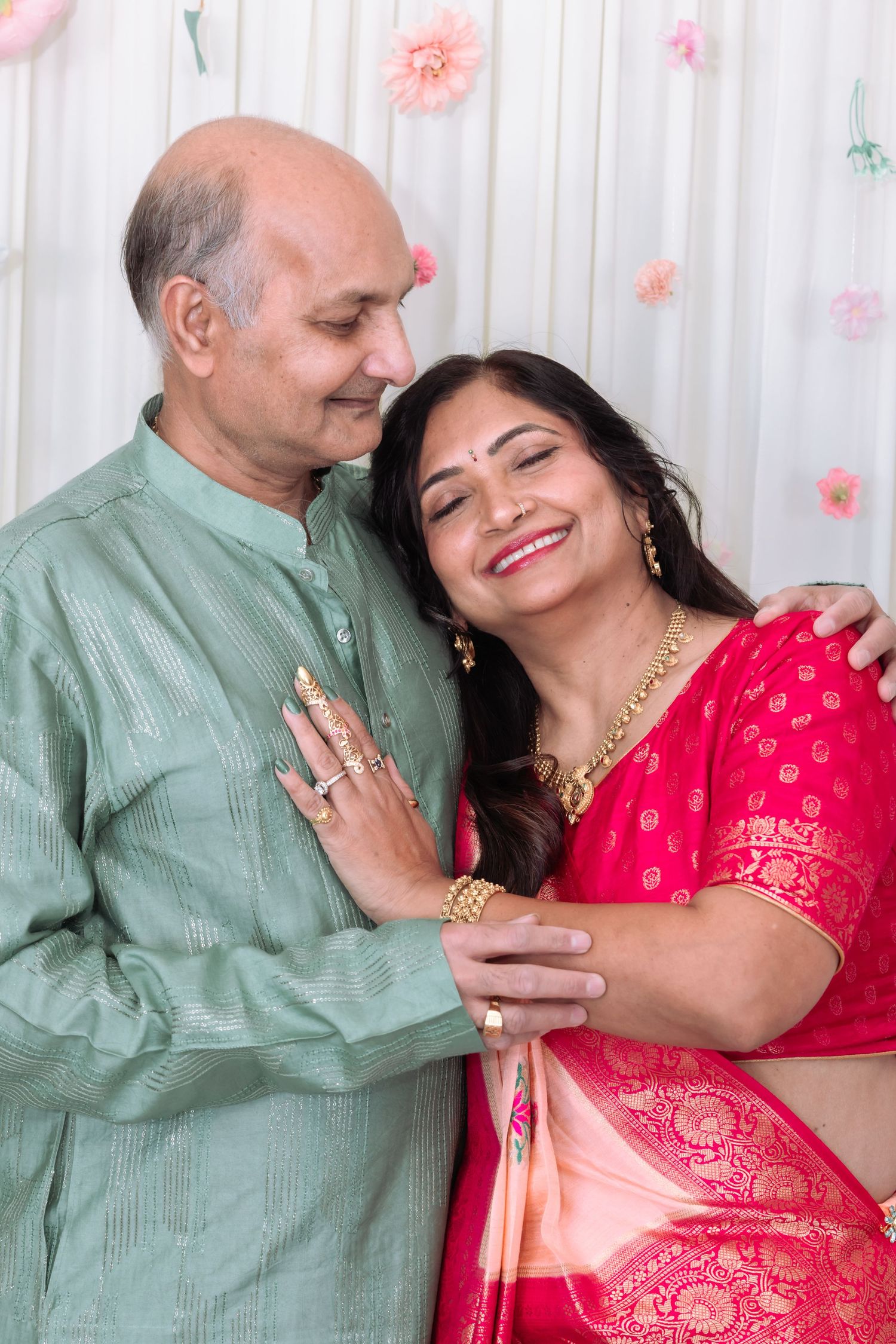 Indian couple in traditional attire embracing, woman in red saree smiling, man in green kurta, floral backdrop.