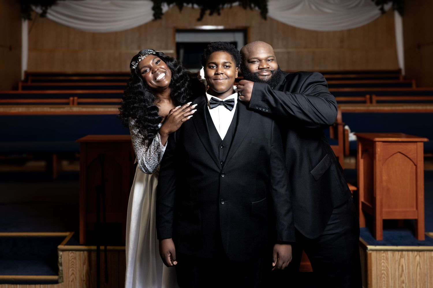Family portrait in a church with a boy in a bow tie tuxedo between two adults in formal attire.