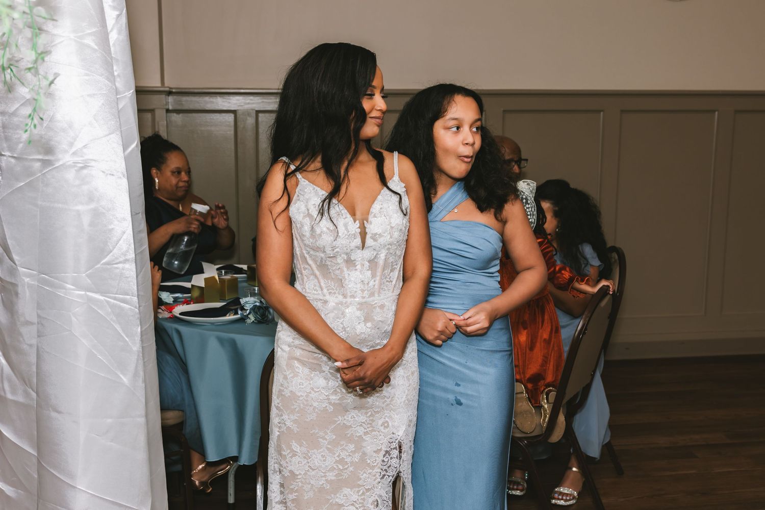 A bride in a white lace gown stands with a bridesmaid in blue at an elegant wedding reception.