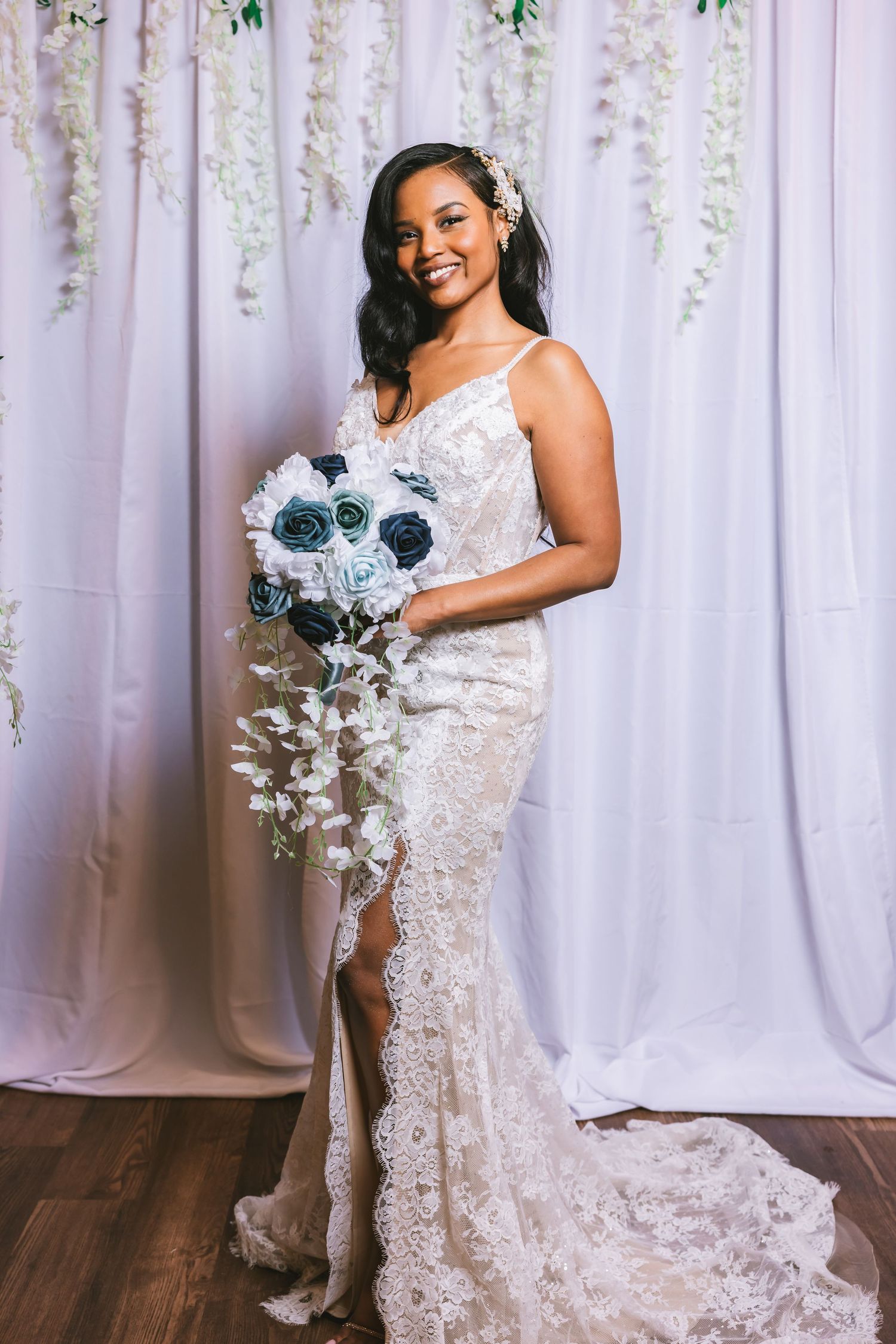 A smiling bride in a lace wedding gown holds a cascading blue and white rose bouquet.