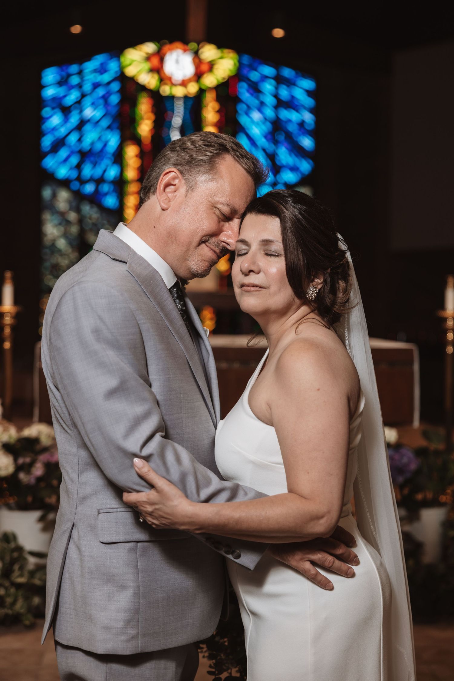 Bride and groom share intimate embrace in church with vibrant blue stained glass window backdrop after wedding ceremony.