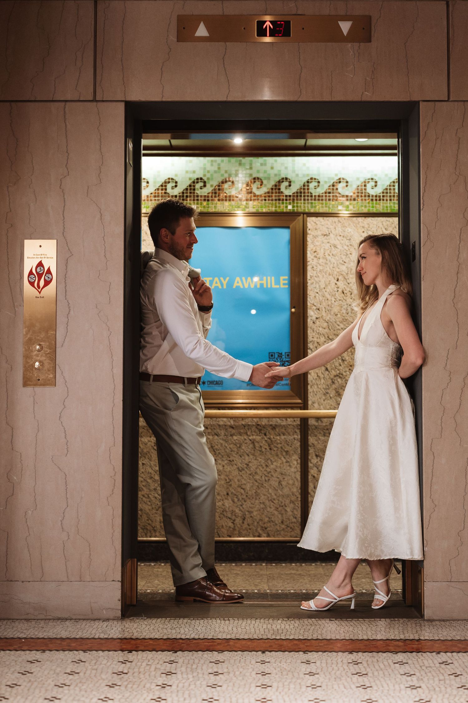 Bride and groom hold hands smiling in an elegant elevator doorway, she in a white midi dress.