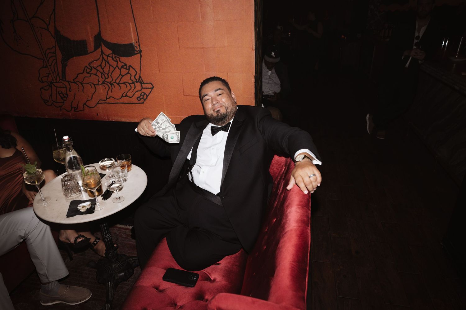 A man in a tuxedo relaxes on a red velvet sofa holding cash at a formal nightclub event.