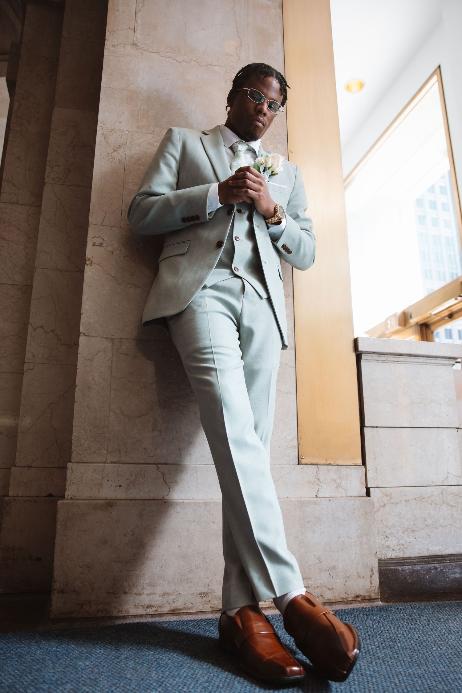 Stylish groom in light gray suit leans against marble column adjusting boutonniere near window.