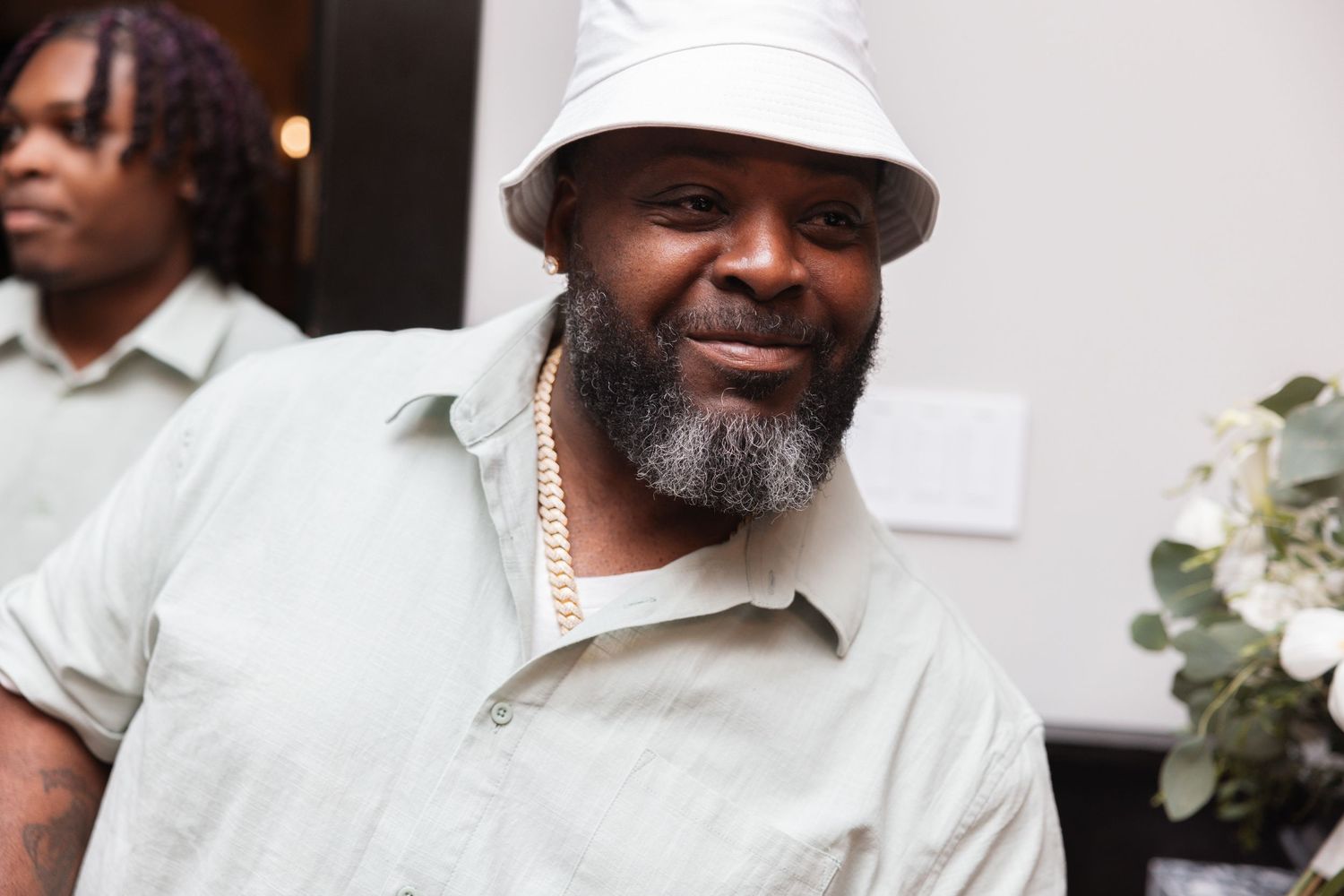 Smiling man in white bucket hat and gold chain poses at wedding celebration event.