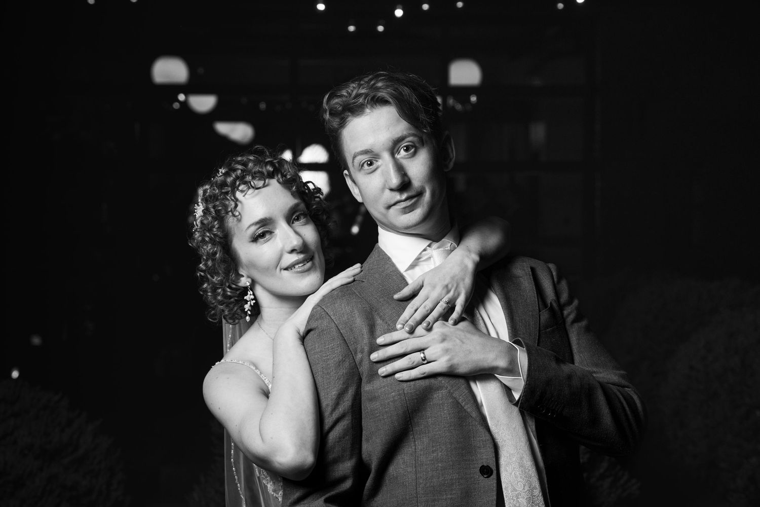 Black and white photo of a couple embracing outdoors at night, smiling warmly under soft string lights.