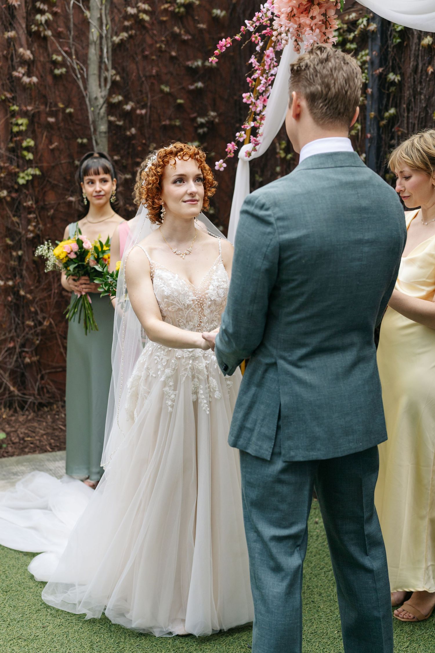 Redhead bride in floral lace gown exchanges vows with groom at outdoor wedding ceremony with floral arch.