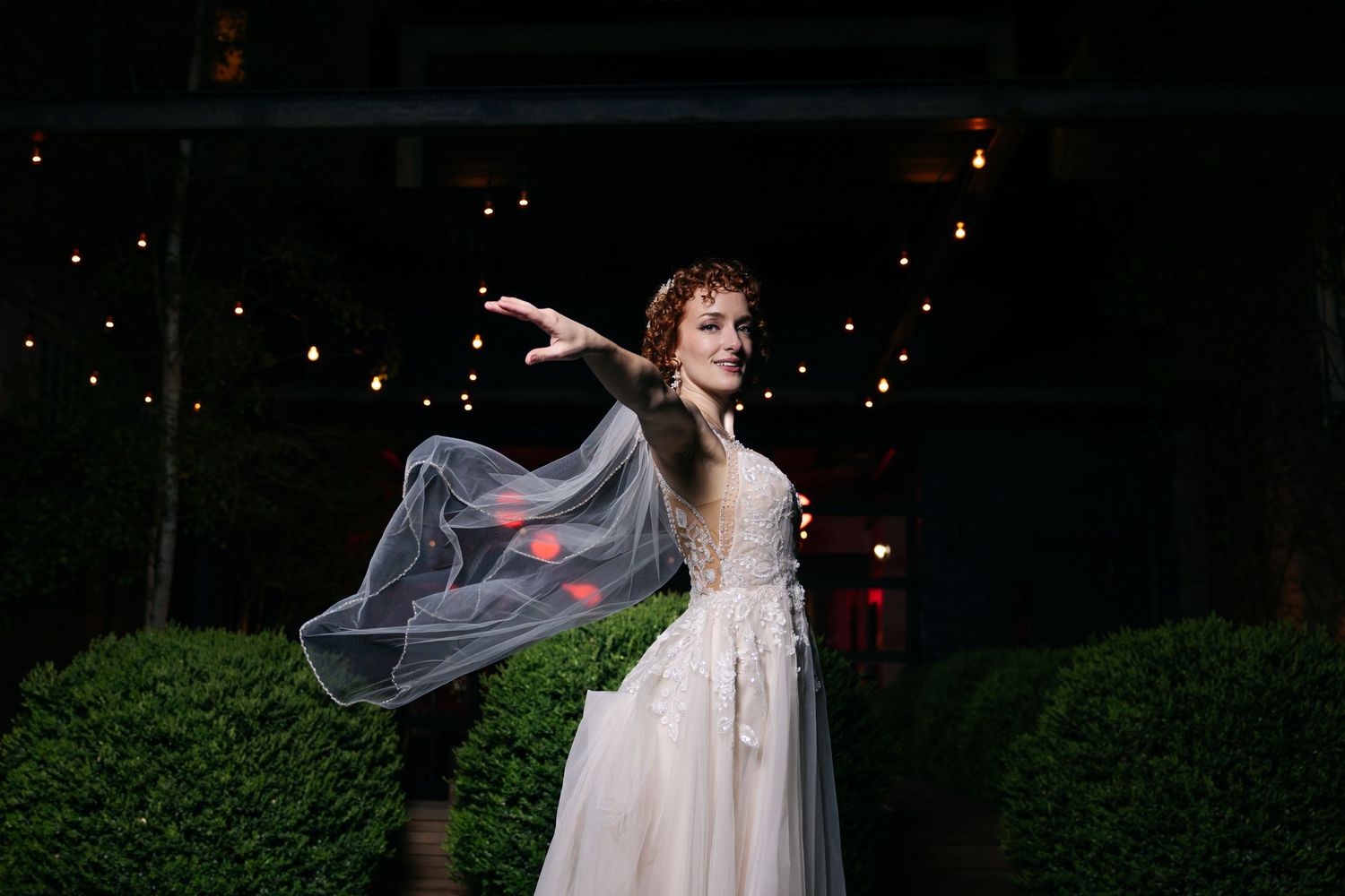Bride in flowing white gown twirling veil at night with string lights glowing in background.