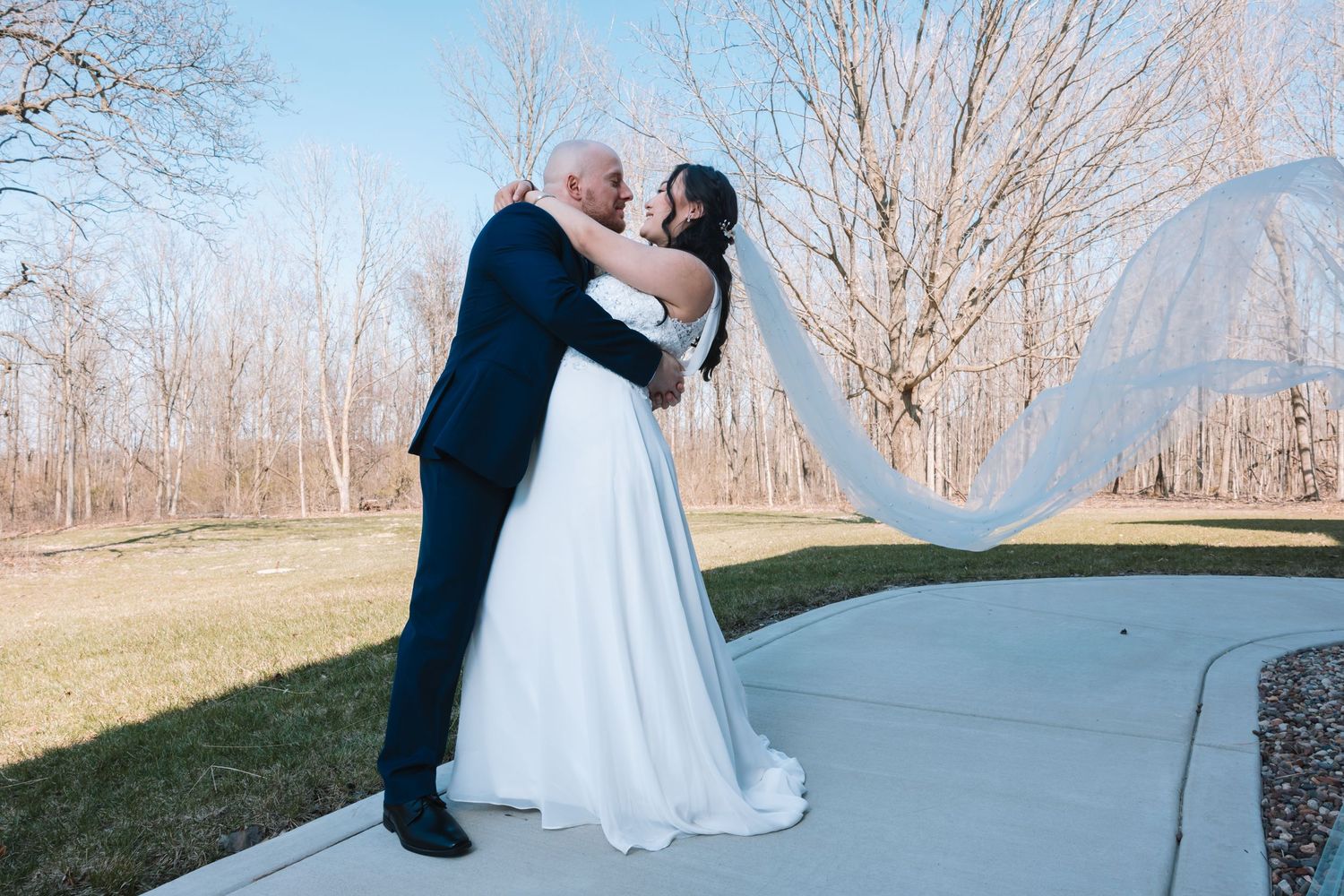 Newlyweds share romantic kiss on sunny outdoor pathway with veil billowing in the wind.