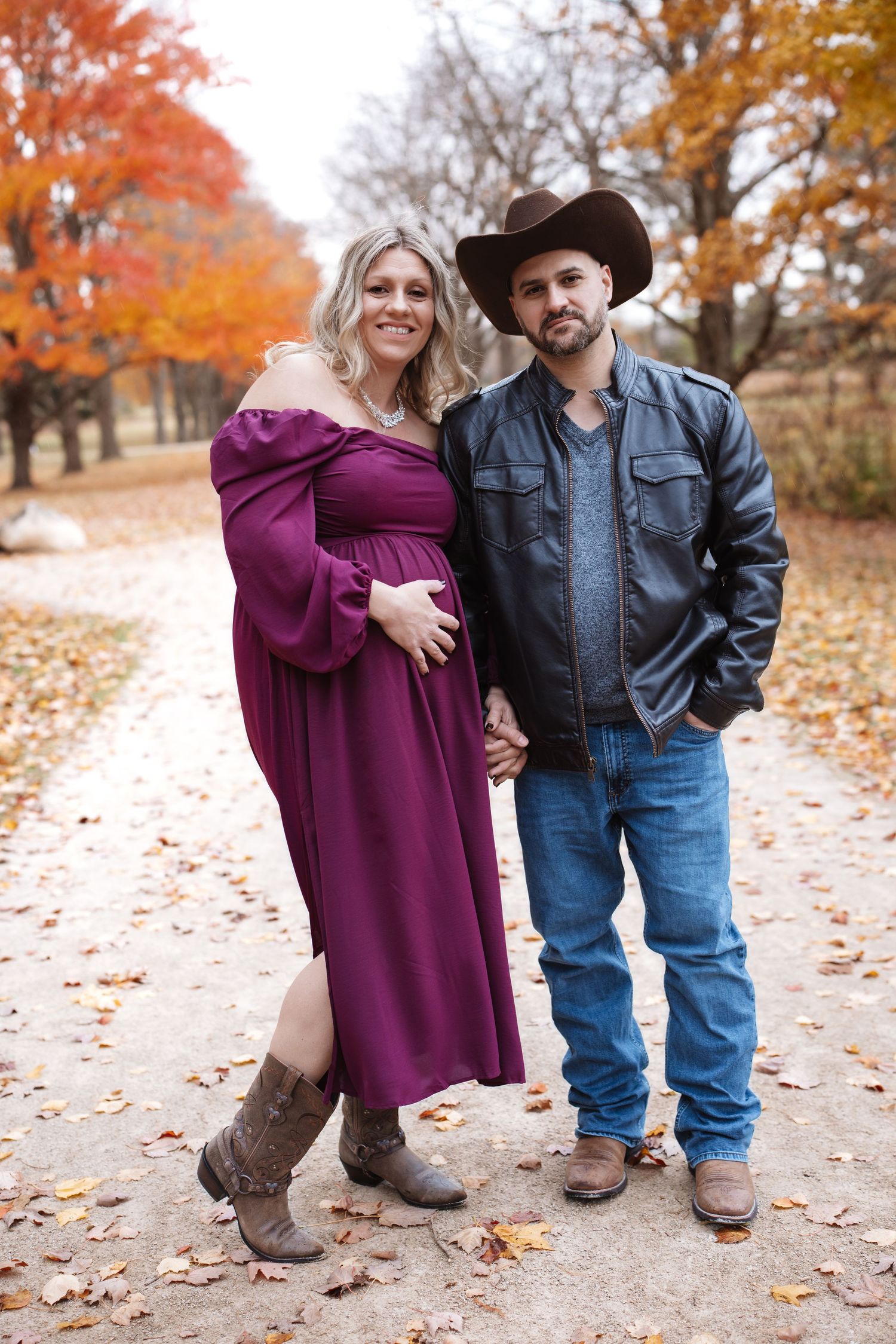 Pregnant woman in burgundy gown poses with partner in cowboy hat on a fall foliage path.