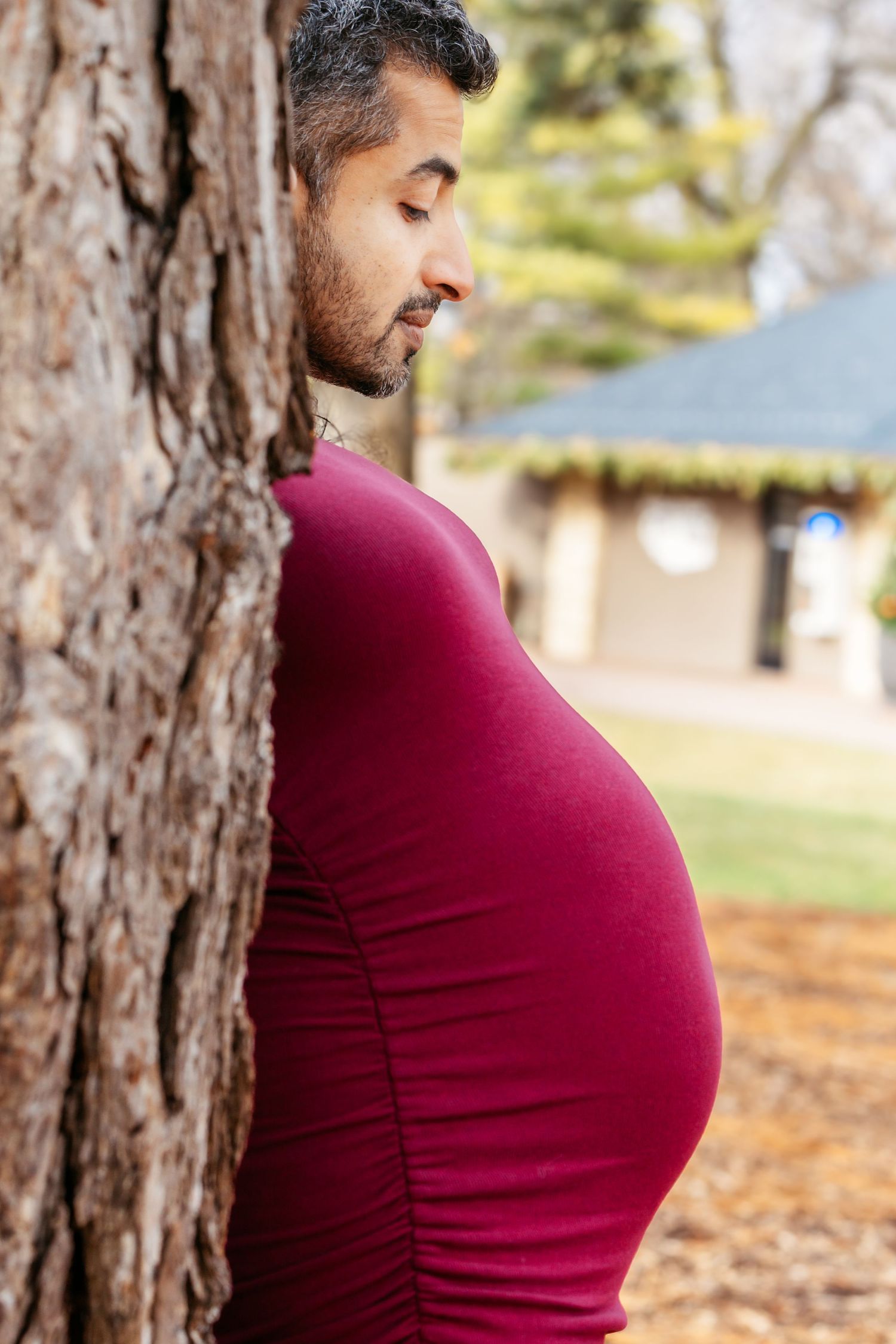 Side profile of pregnant woman in red dress leaning against a tree trunk in a park.