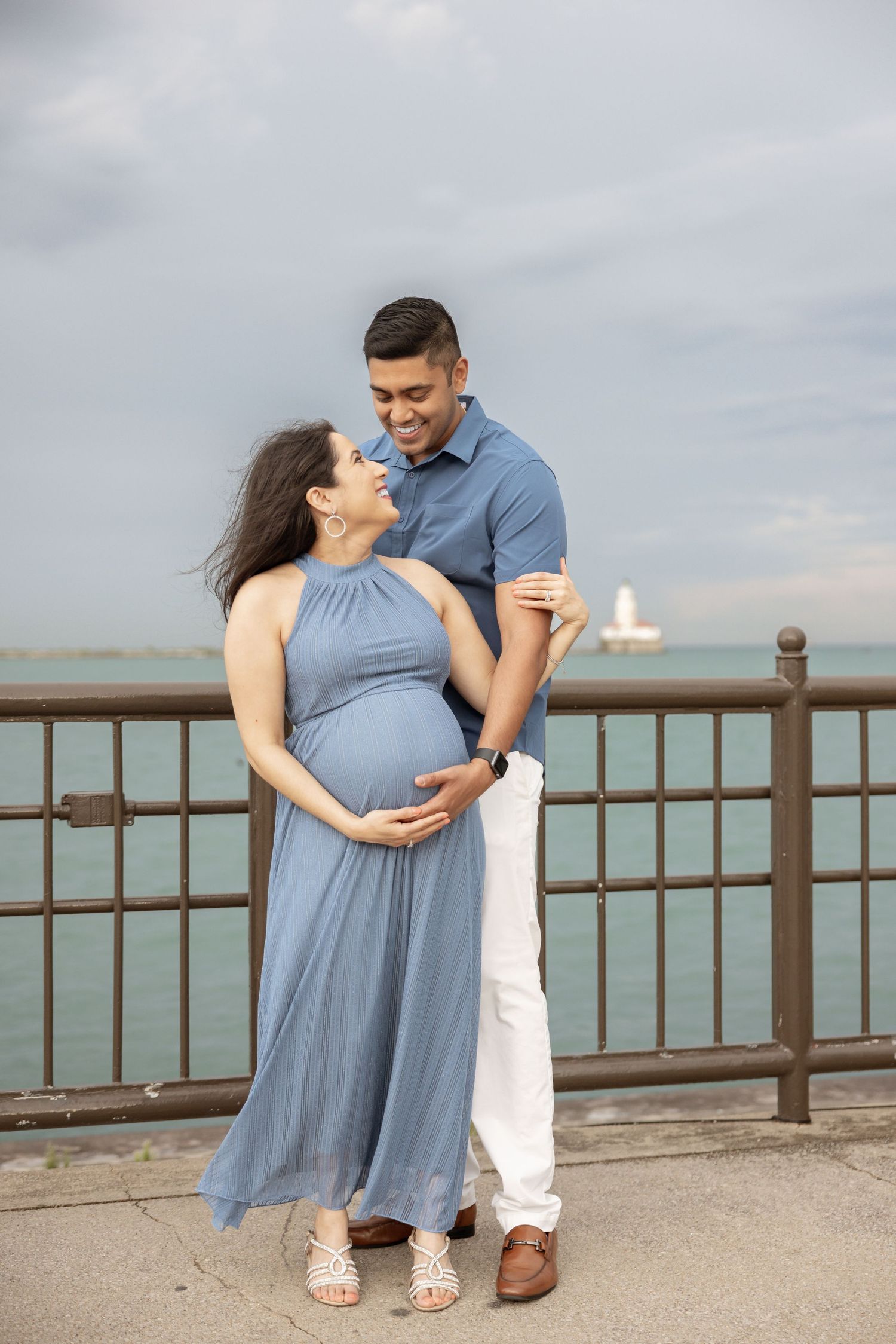 Joyful pregnant couple in coordinating blue outfits embracing and smiling by a waterfront railing.
