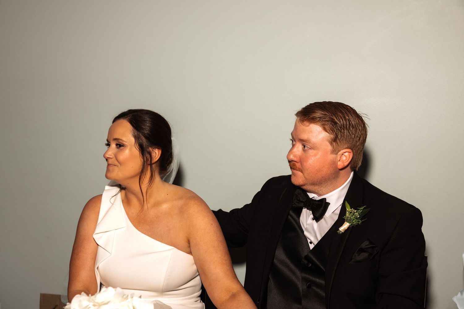 Bride in white one-shoulder gown sits with groom in tuxedo who rests hand on her shoulder at wedding.