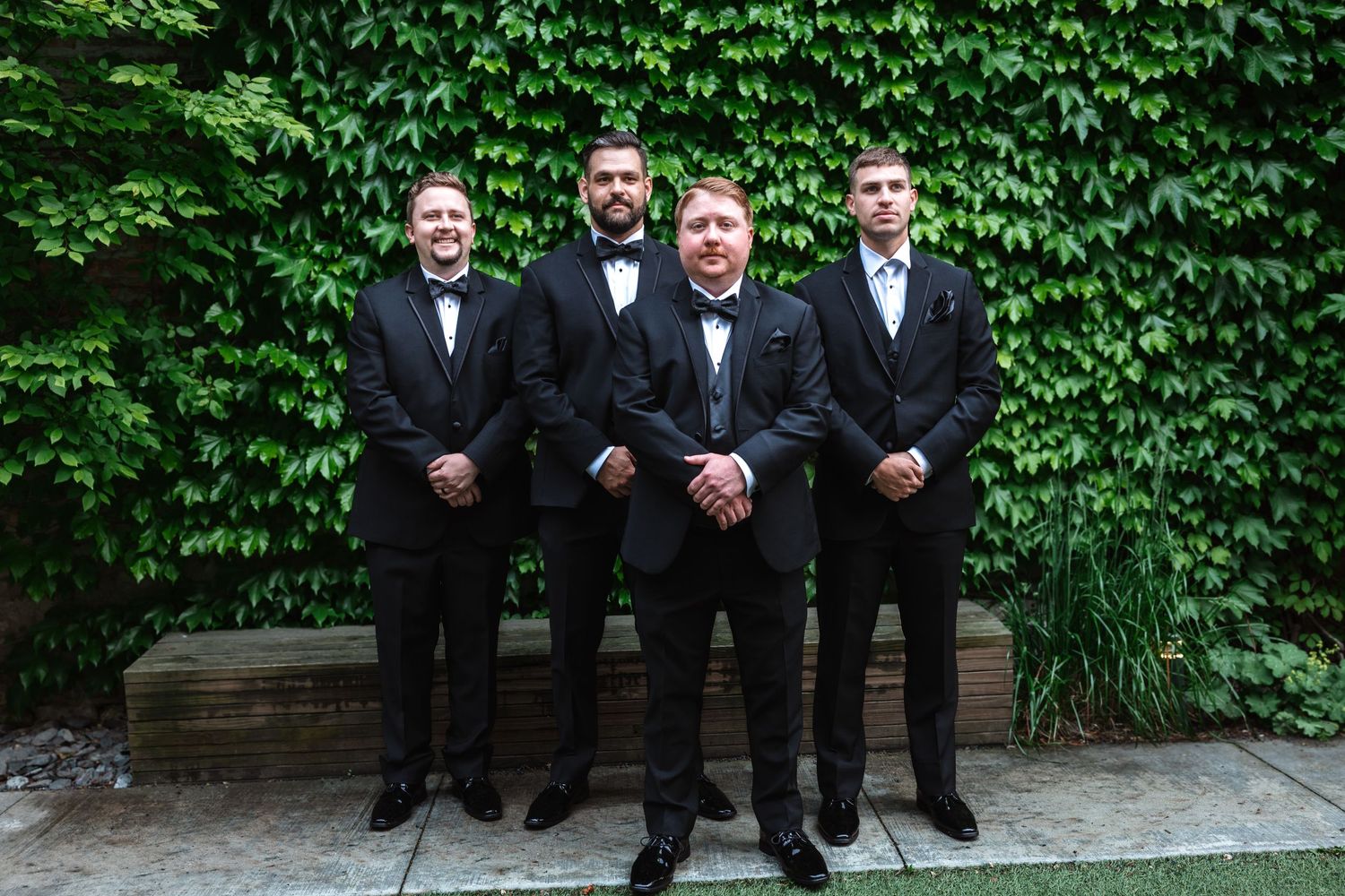 Four groomsmen in black tuxedos posing seriously in front of a green ivy-covered wall.