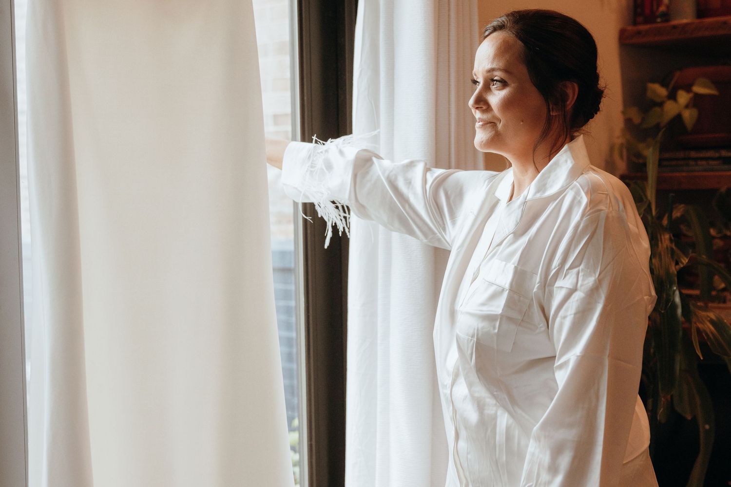 Bride in feather-trimmed white robe looking thoughtfully out window on her wedding morning.