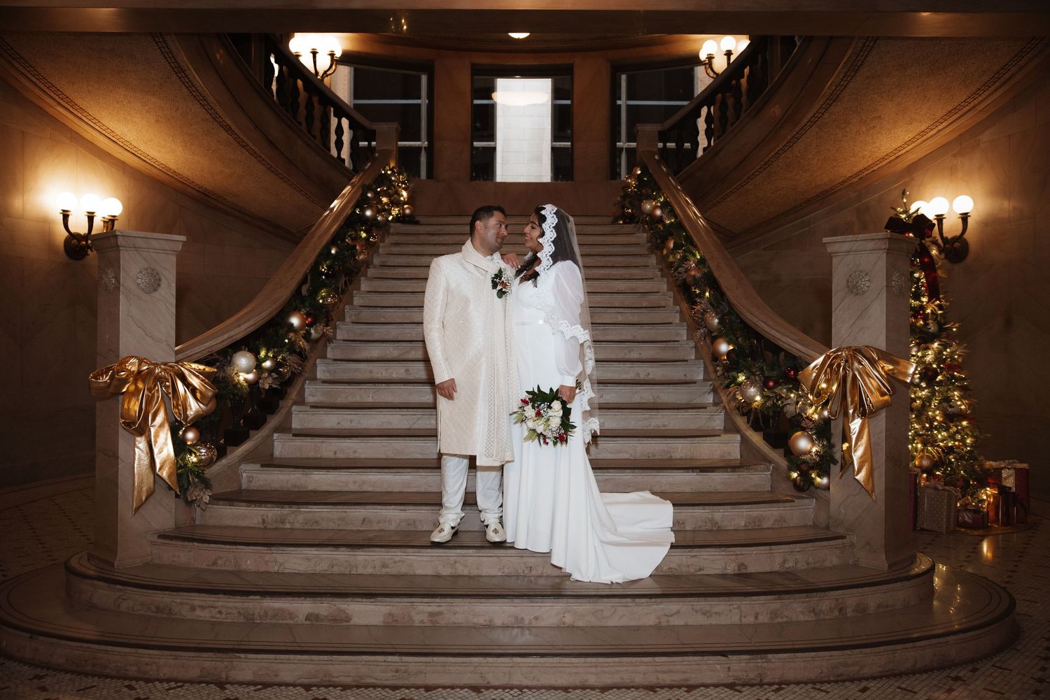 Bride and groom sharing intimate moment on grand staircase with Christmas garland and golden bow decorations.