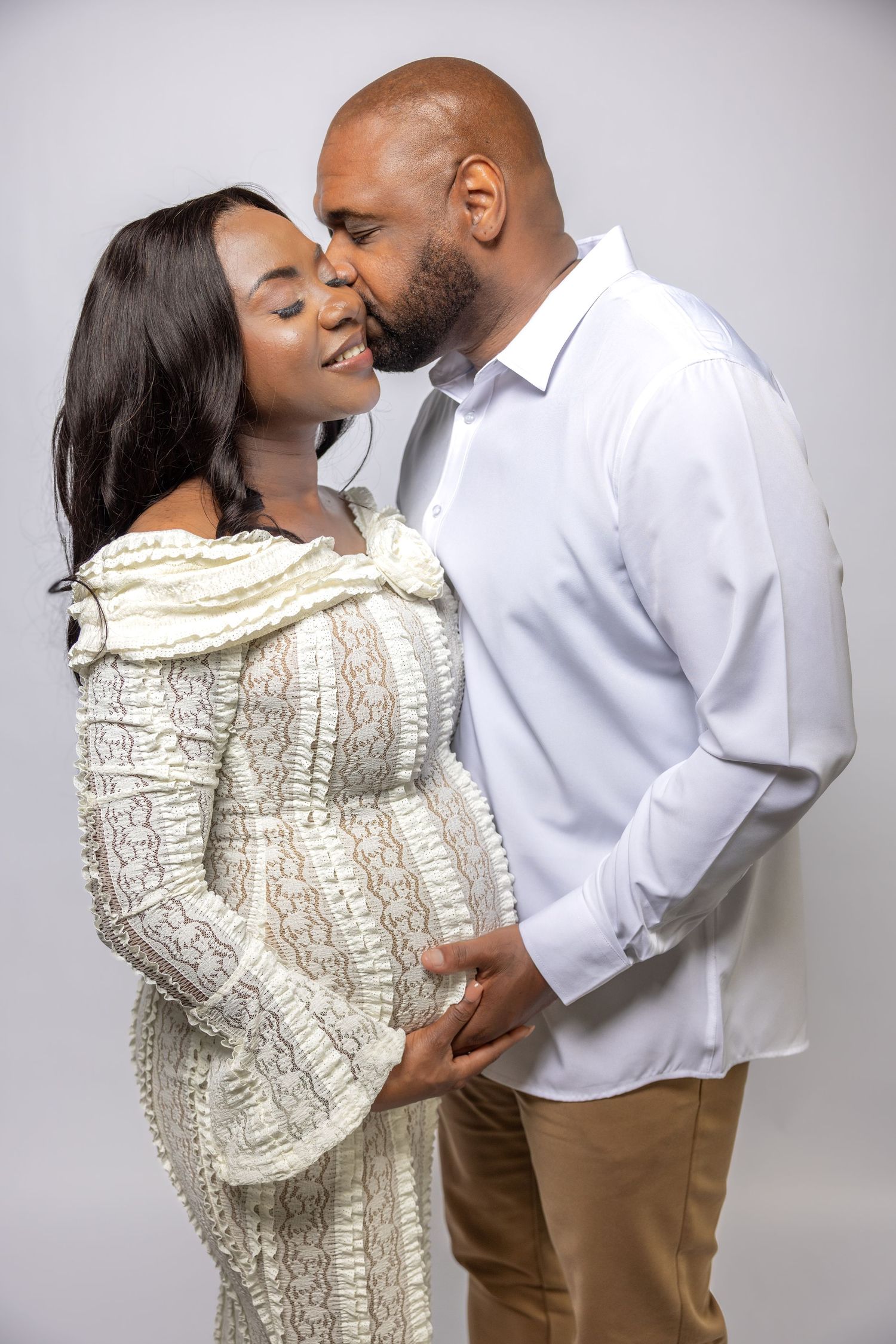 Man kissing pregnant woman's cheek in studio, woman in white lace dress holding baby bump.