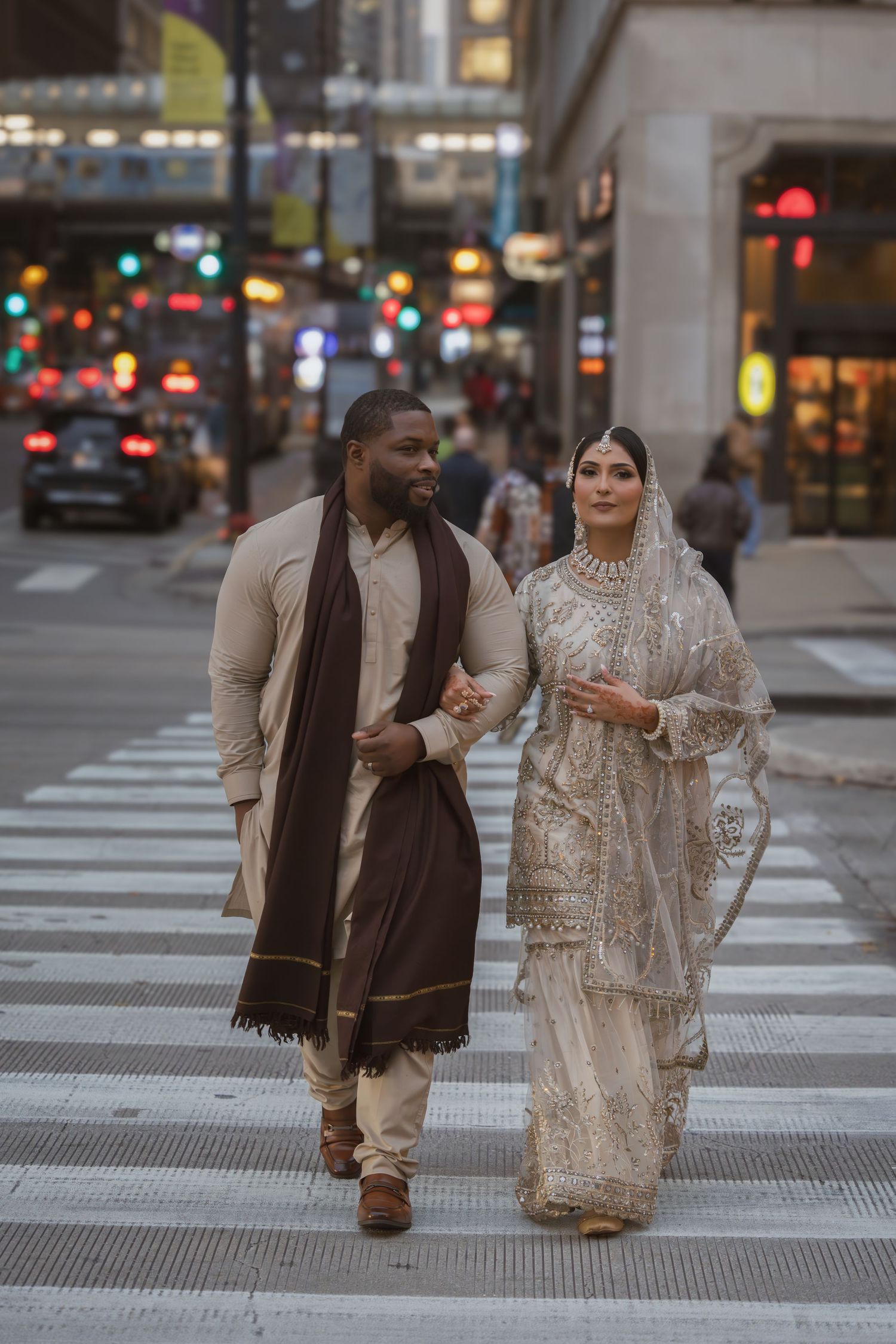 Bride in ivory embroidered lehenga and groom in beige sherwani crossing a busy urban street together.