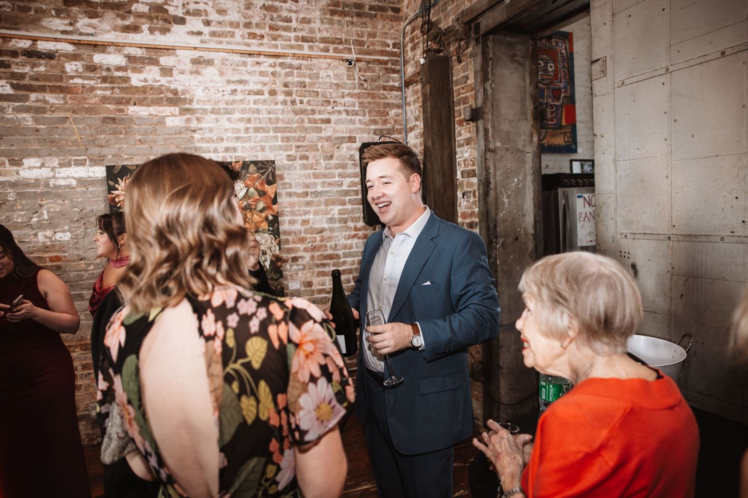 Man in blue blazer laughing with guests at a cocktail reception near brick wall.