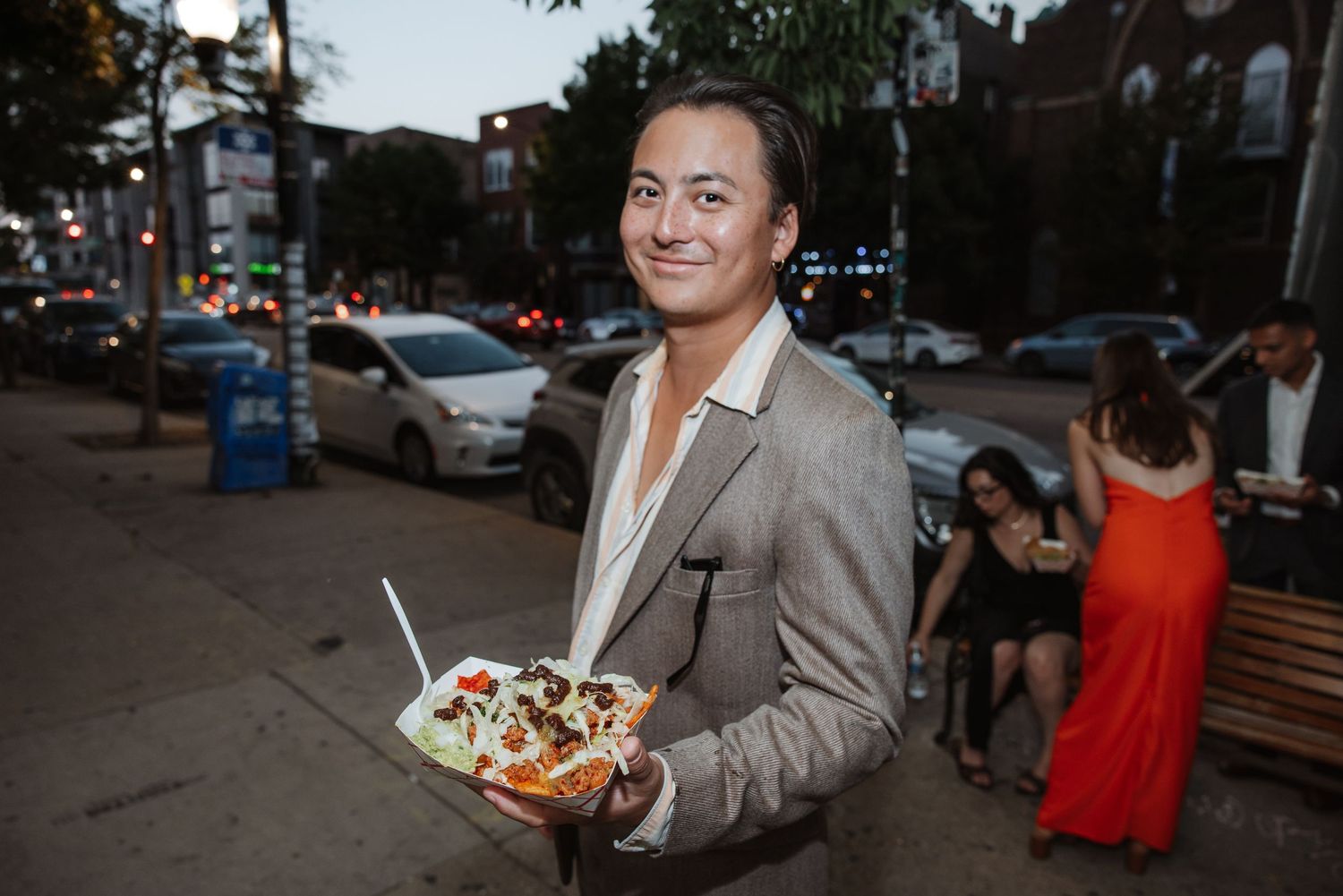 Woman in blazer smiling while holding a loaded street food dish at an outdoor evening gathering.