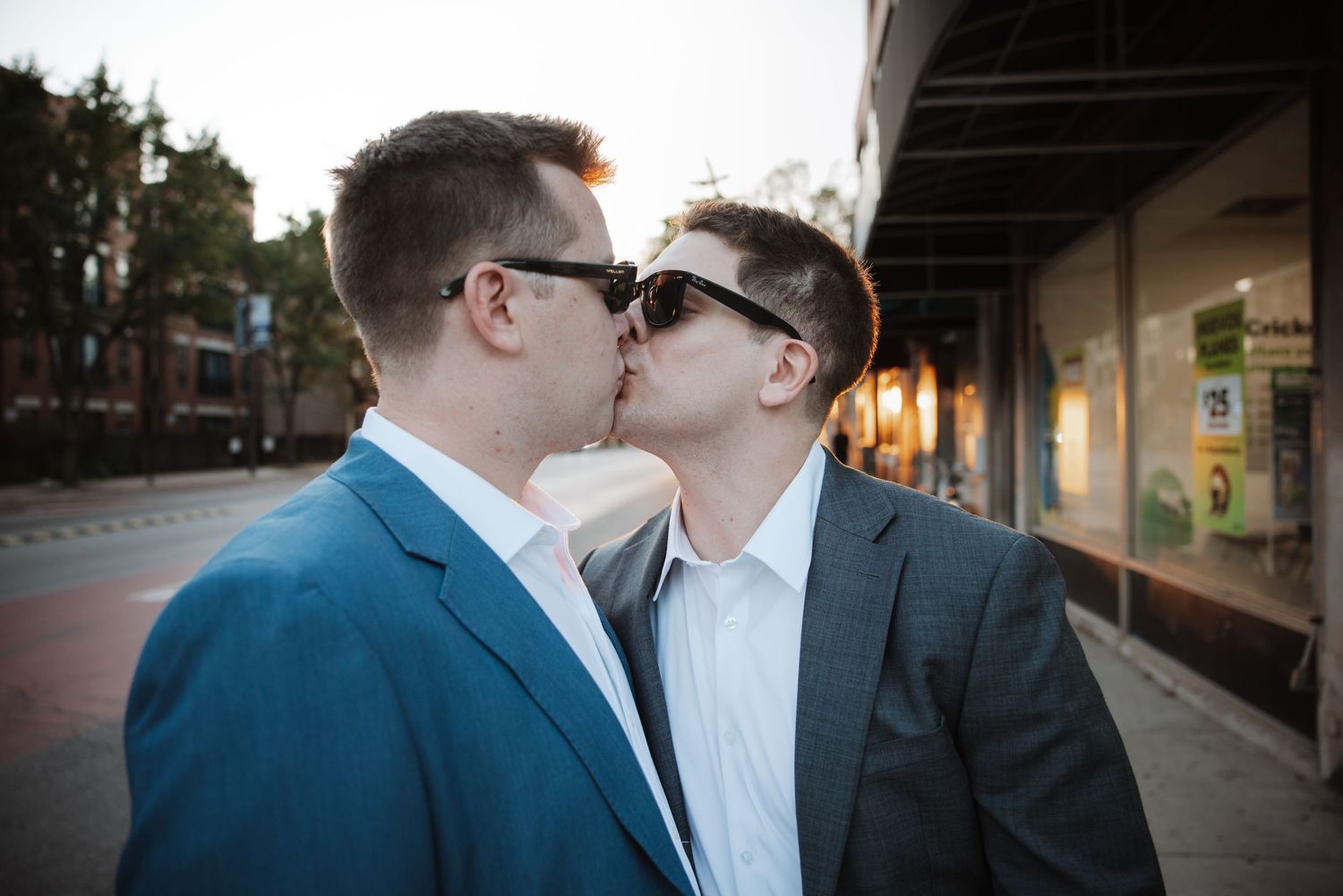 Two grooms in suits sharing a kiss on a city sidewalk during their wedding celebration.