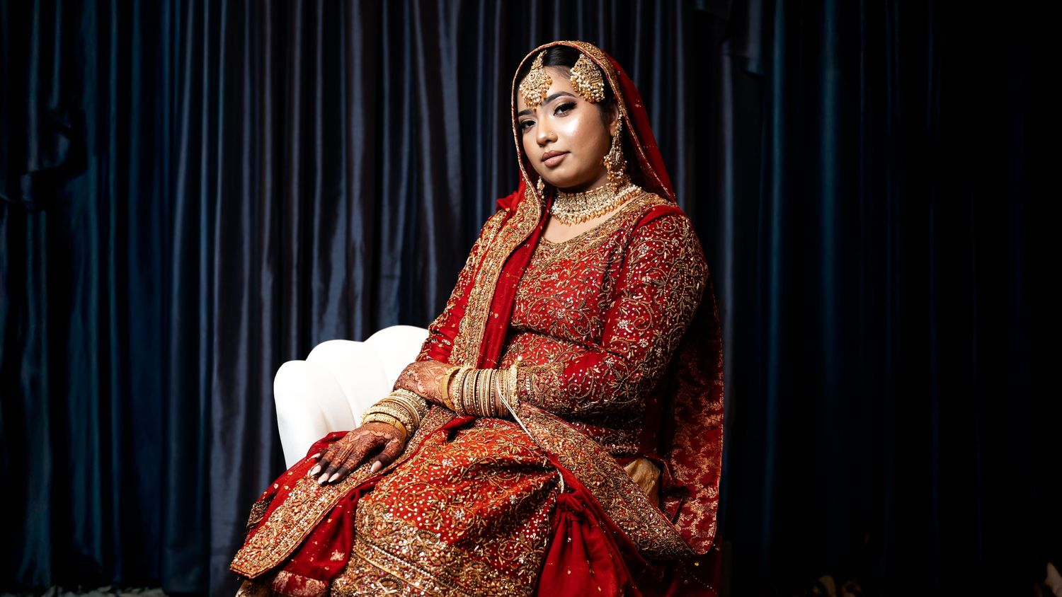 Bride in red embroidered South Asian bridal outfit seated against blue curtain backdrop.