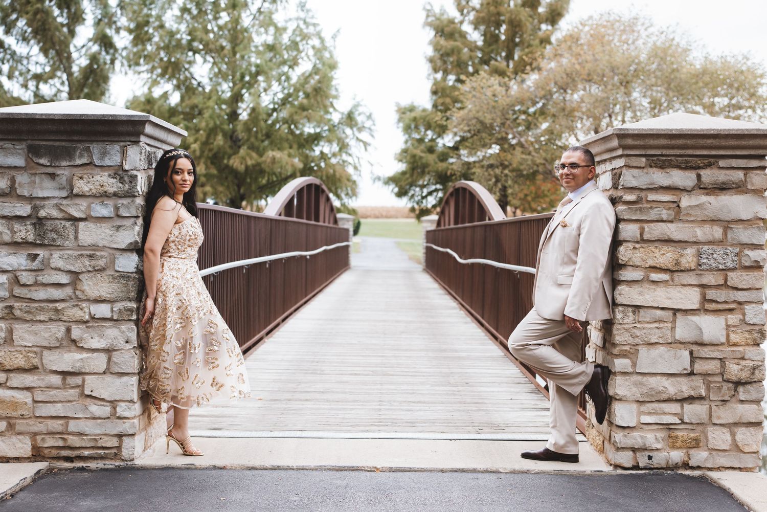 Wedding couple posing at opposite sides of stone bridge entrance in elegant attire.