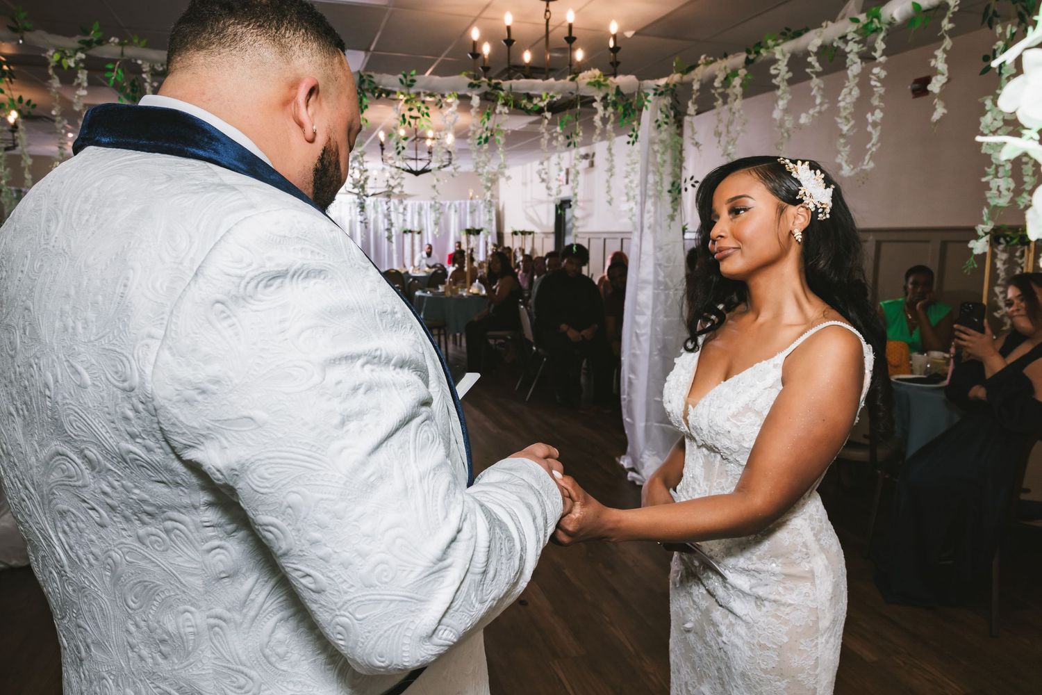 Bride and groom hold hands while exchanging heartfelt wedding vows at romantic indoor ceremony with floral decor.