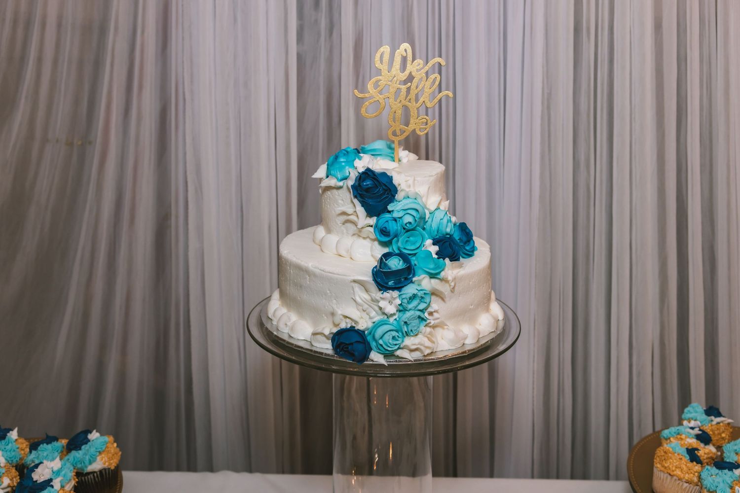 Three-tier white wedding cake decorated with blue and teal flowers and a gold monogram topper on a clear stand.