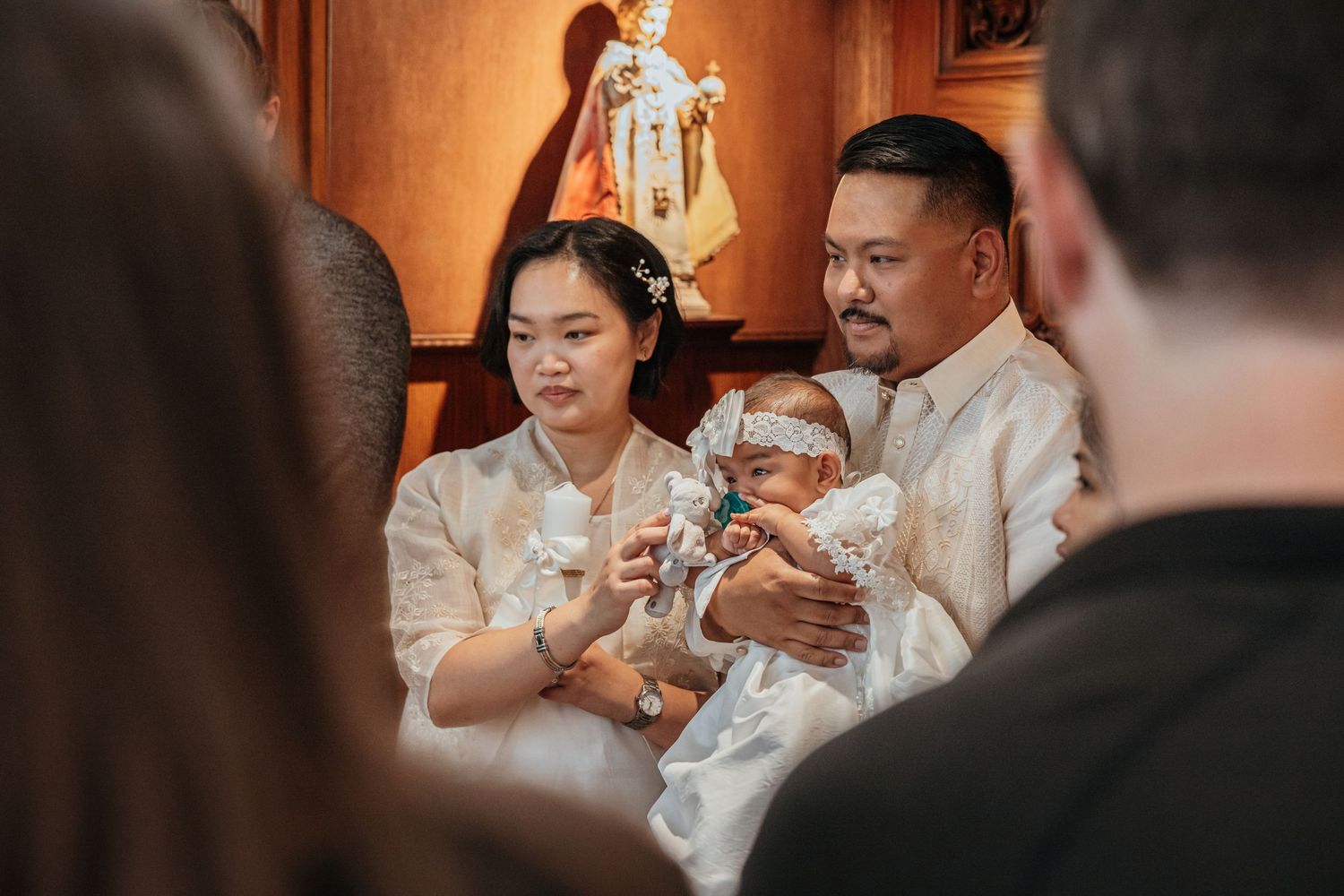 Filipino parents in matching white barong attire tenderly hold their infant during a Catholic baptism.