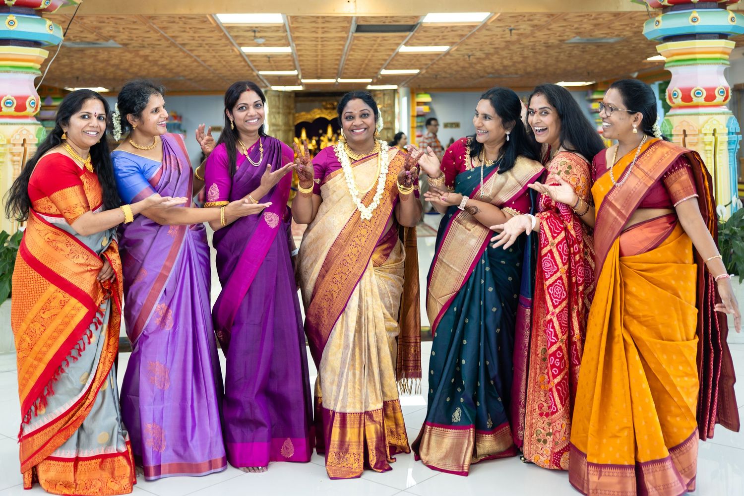 Seven women in vibrant silk sarees laughing and posing together inside a decorated Hindu temple.