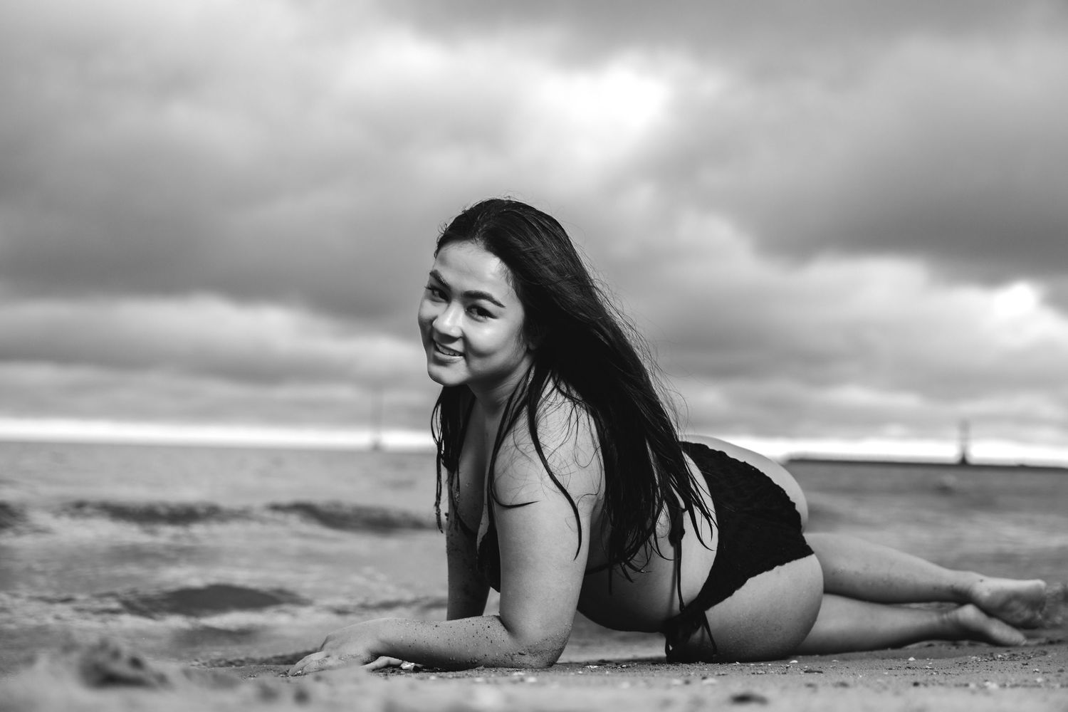 Black and white photo of woman in bikini lying on sandy beach with dramatic cloudy sky.