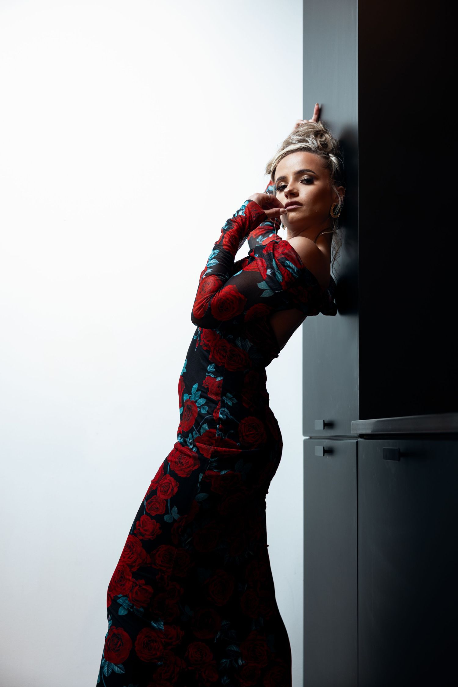 Woman in red floral maxi dress leaning against dark wall in elegant fashion pose.